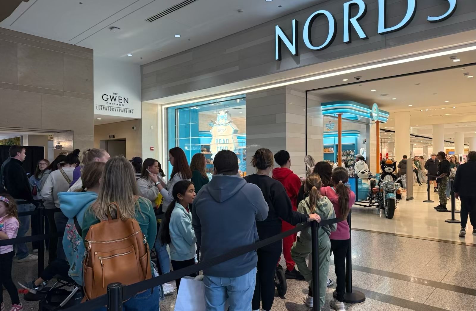 Crowd of people, including children and adults, queued inside a mall near a large store entrance with partial signage "NORDSTROM" and a blue-themed display featuring plush toys and a motorbike inside.