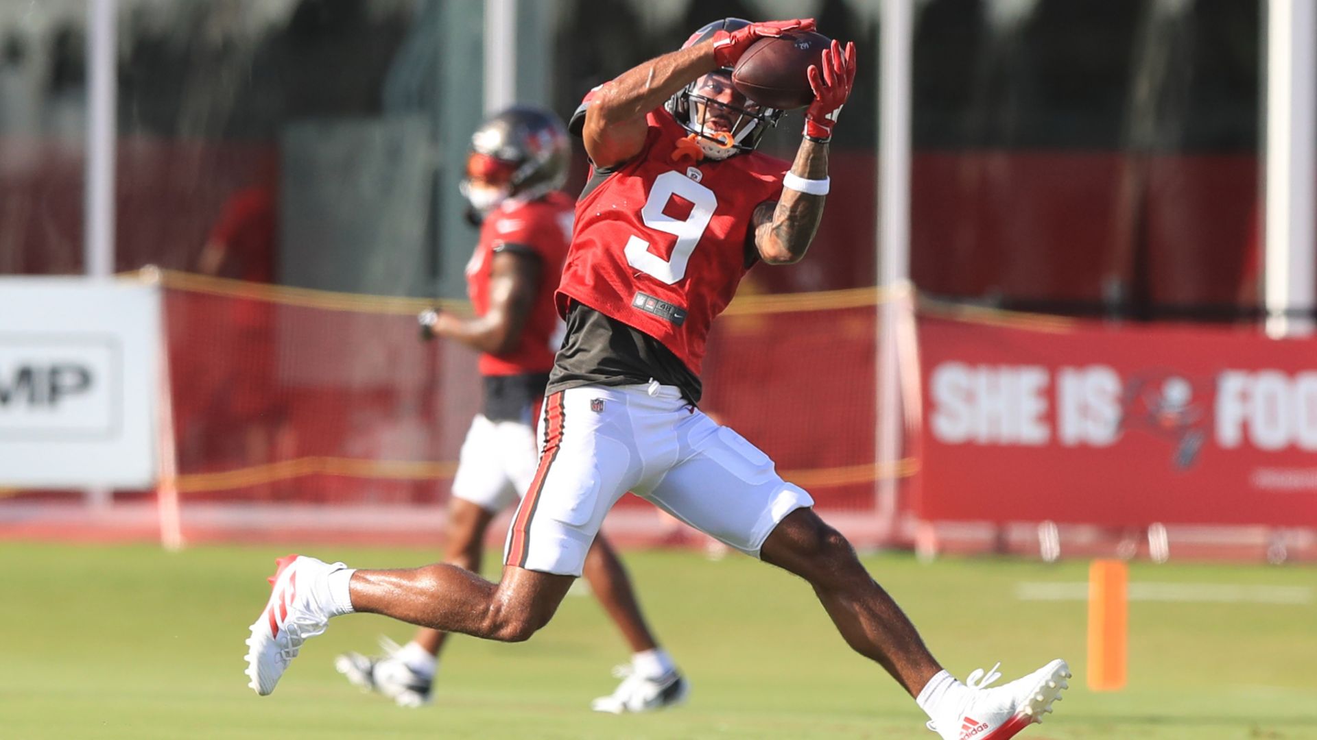 Football player in red jersey number 9 and white pants catching a football mid-air on a green field during practice, with another player and a red banner in the background.