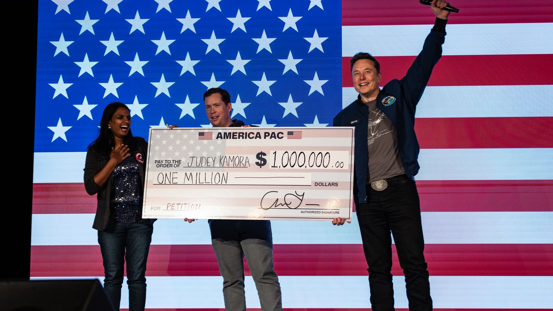 Elon Musk awards a person named Judey Kamora with a $1,000,000 check during an America PAC town hall in front of an American flag.
