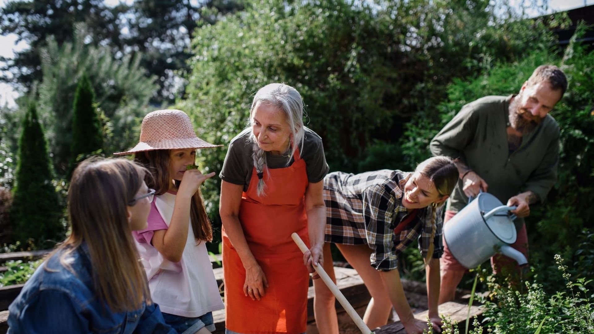 Outdoor garden scene with several people gathered around garden beds; a gray-haired woman in an orange apron guides others, while a bearded man waters plants with a large bucket amid lush greenery.