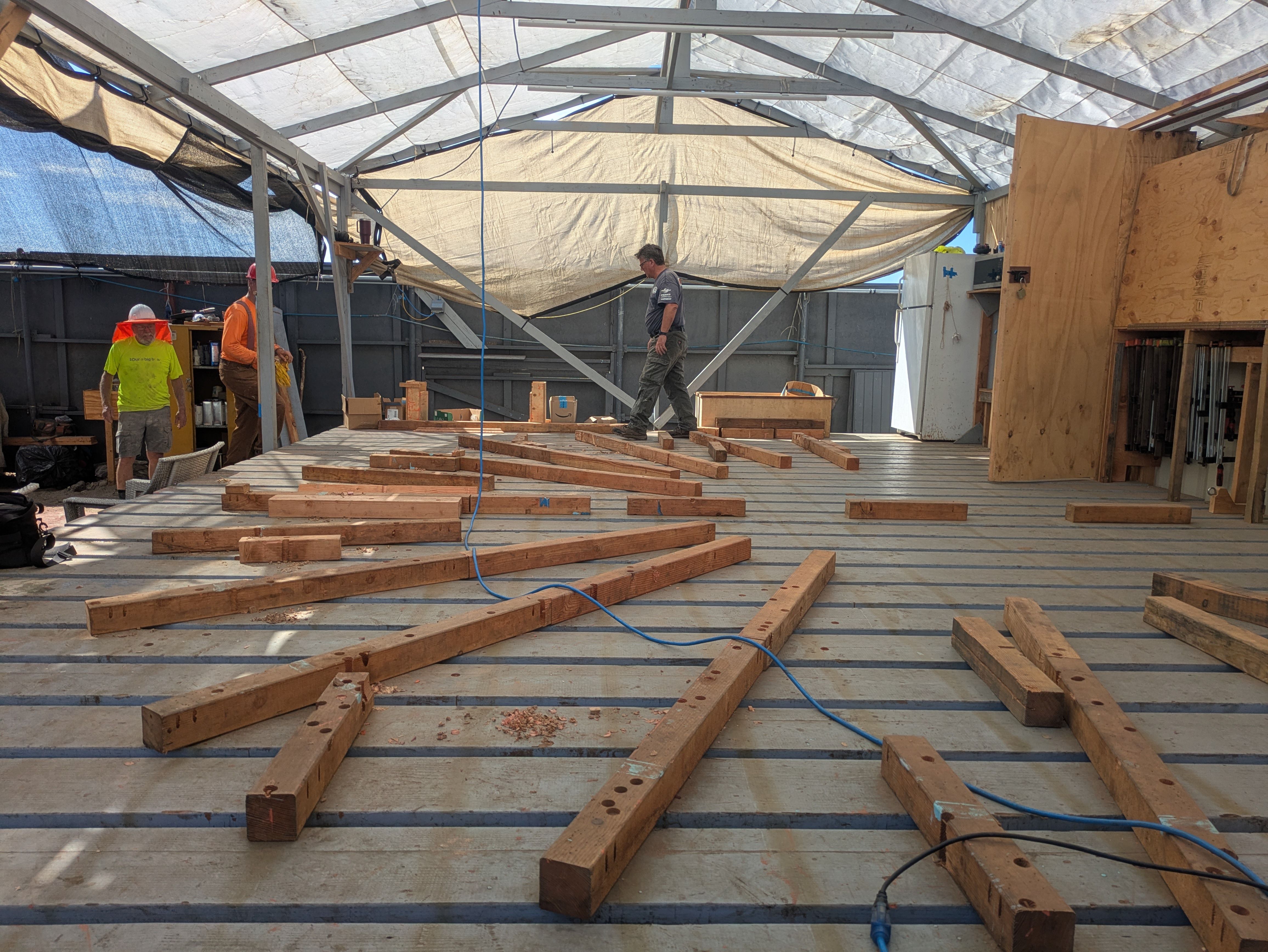 Inside a makeshift shelter with a metal frame and beige tarp roof, three workers stand among scattered lumber planks and tools, plywood walls, and shelves along the sides.