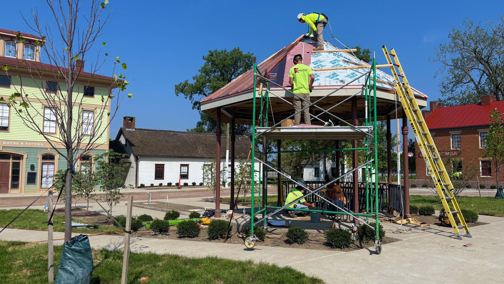 Two workers in neon shirts stand on scaffolding atop a gazebo-like pavilion, installing copper roofing. A yellow ladder leans nearby; blue sky and colorful storefronts provide a bright backdrop.