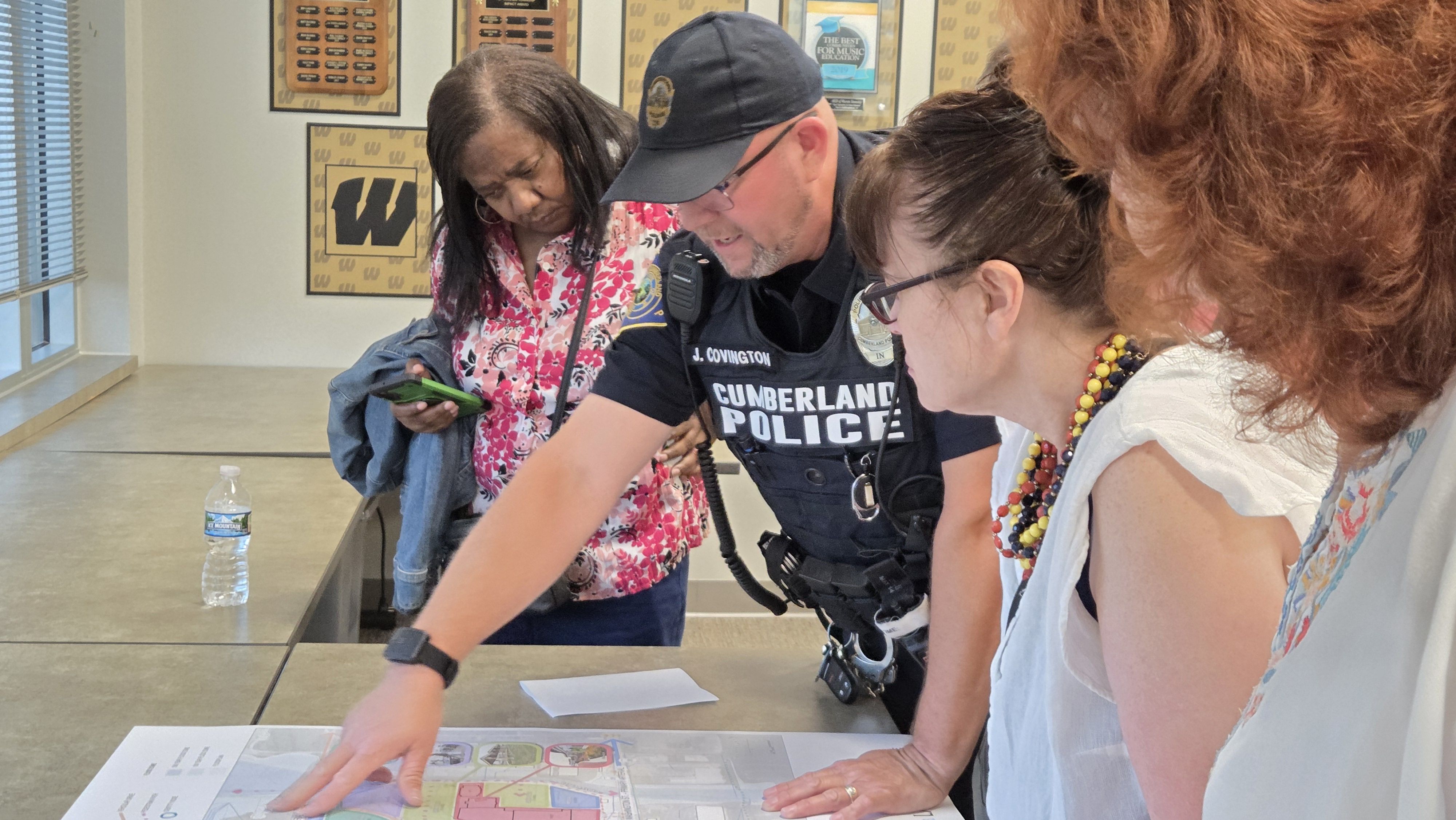Four people lean over a table, studying a large colored map. A Cumberland Police officer in uniform joins two women and a red-haired person as they point to papers.