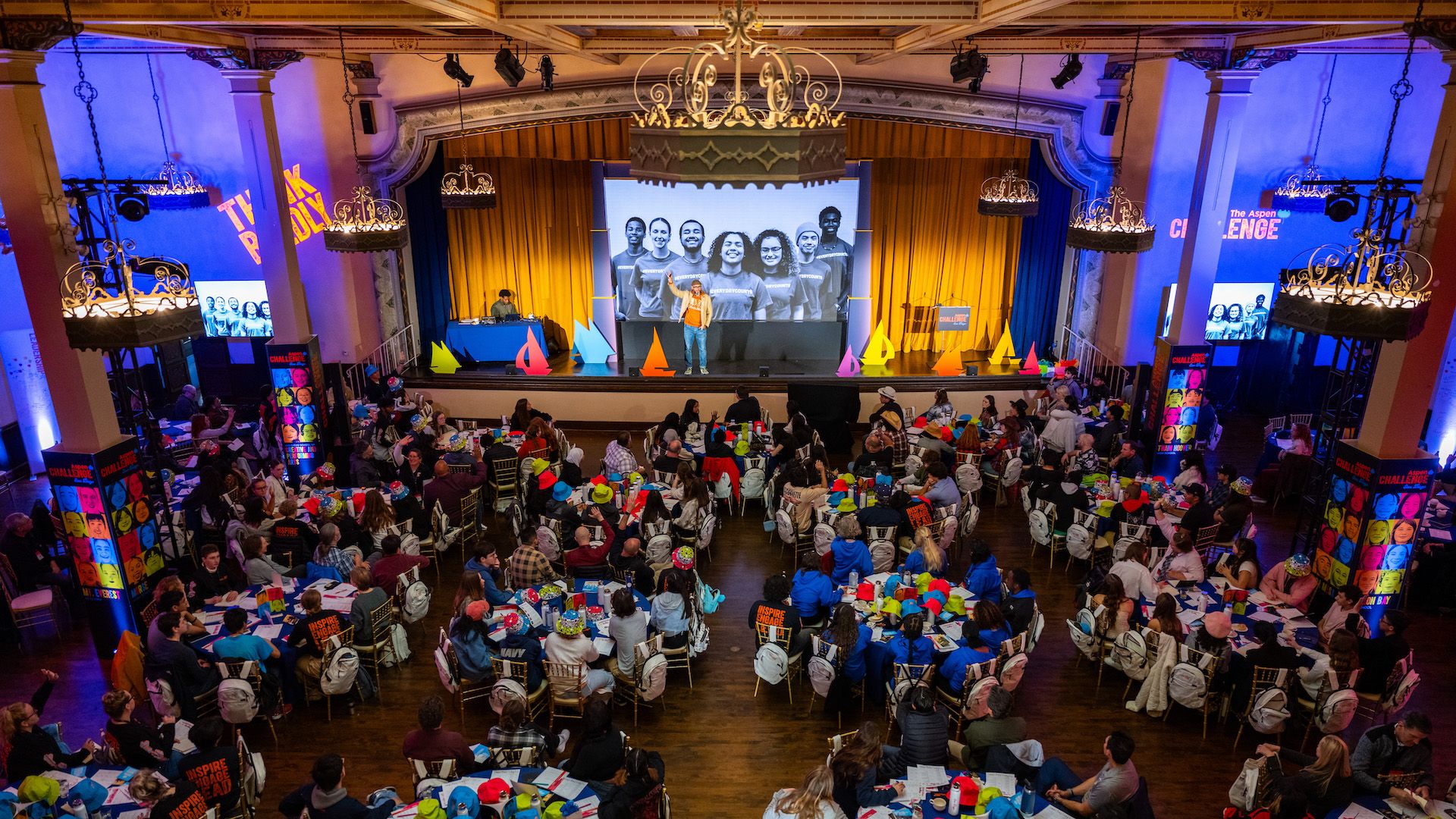 More than 100 students sit at tables looking towards a stage in a ballroom. 