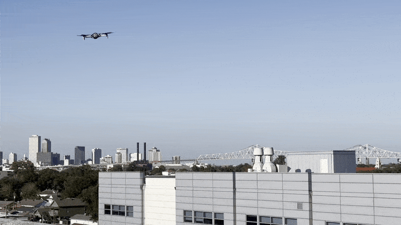 Drone flying above buildings with a city skyline and a large bridge in the background under a clear blue sky.