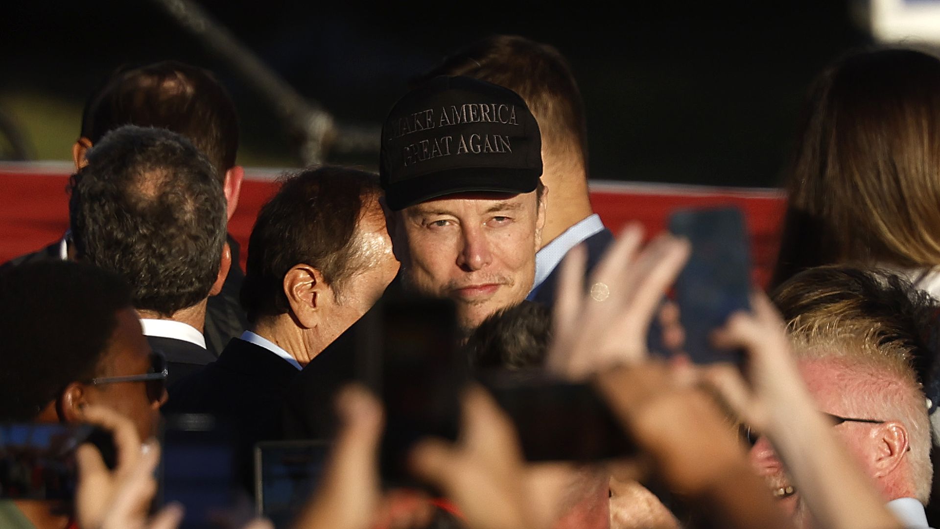 A man in a "make america great again" hat smirking and looking onto the crowd, which is blurred around him