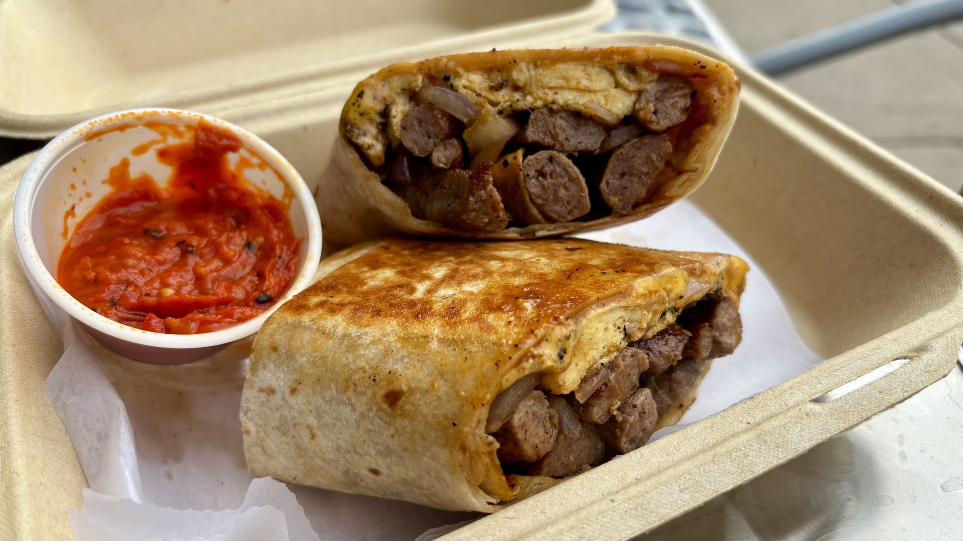 Grilled wrap cut in half with meat and eggs, served in a takeout box with a side of red sauce.
