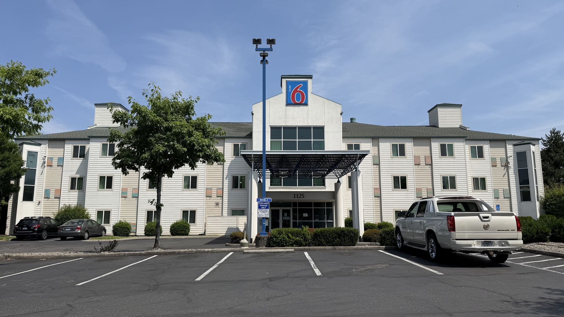 A white three-story Motel 6 building in North Portland with a large blue and red sign, flanked by trees and a nearly empty parking lot under a clear blue sky.