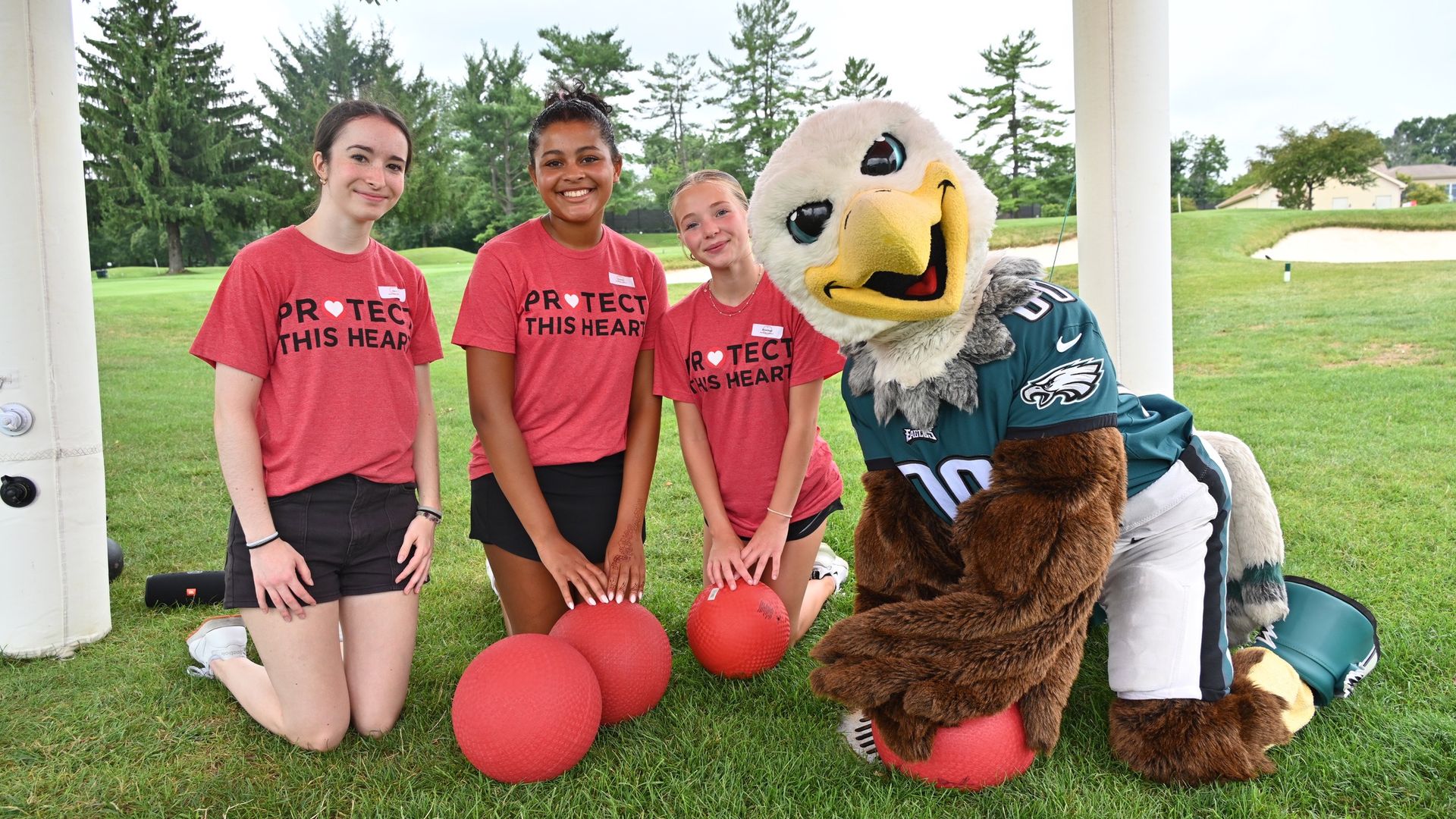 Three smiling girls in red t-shirts stand beside Eagles mascot Swoop in a teal jersey on a grassy field; red balls sit in the foreground, under a pavilion with trees in the background.