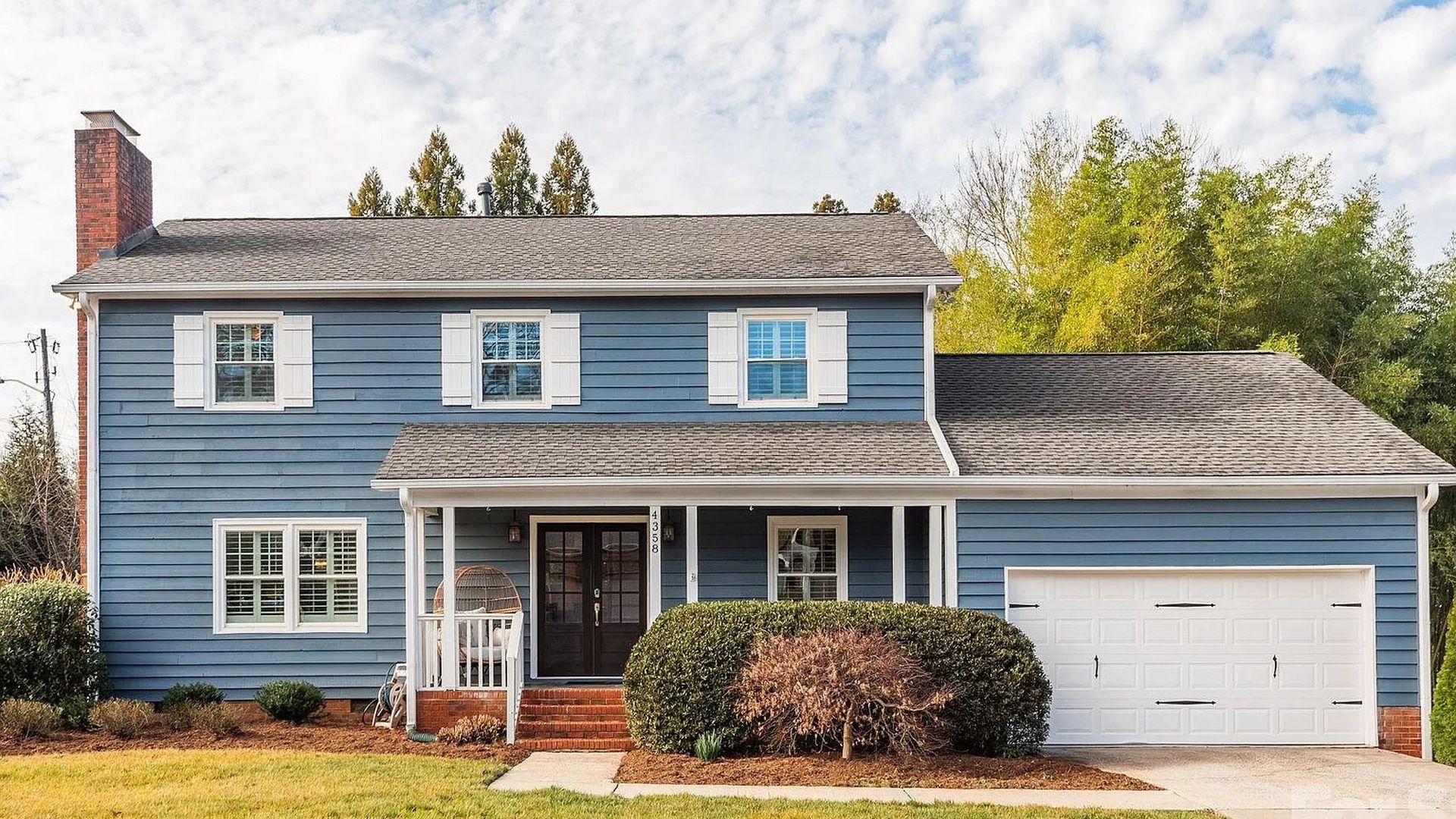 Two-story blue house with white shutters, a brick chimney, and a white double garage door. Front porch with seating, shrubs, a small tree, and a lawn under a partly cloudy sky.