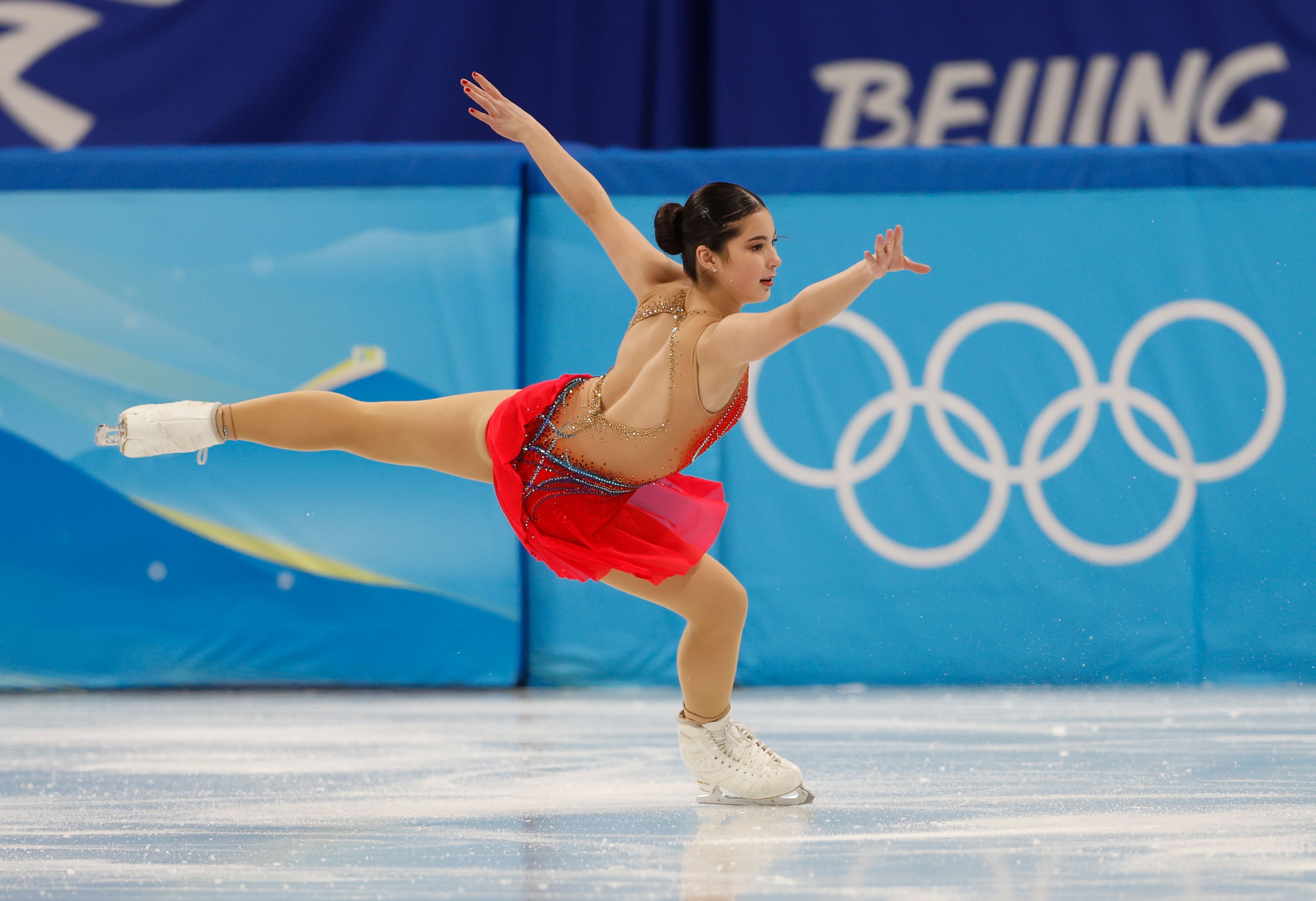 Female figure skater in a red dress performing a spin on ice with one leg extended, Olympic rings and "Beijing" text visible in the blue background.