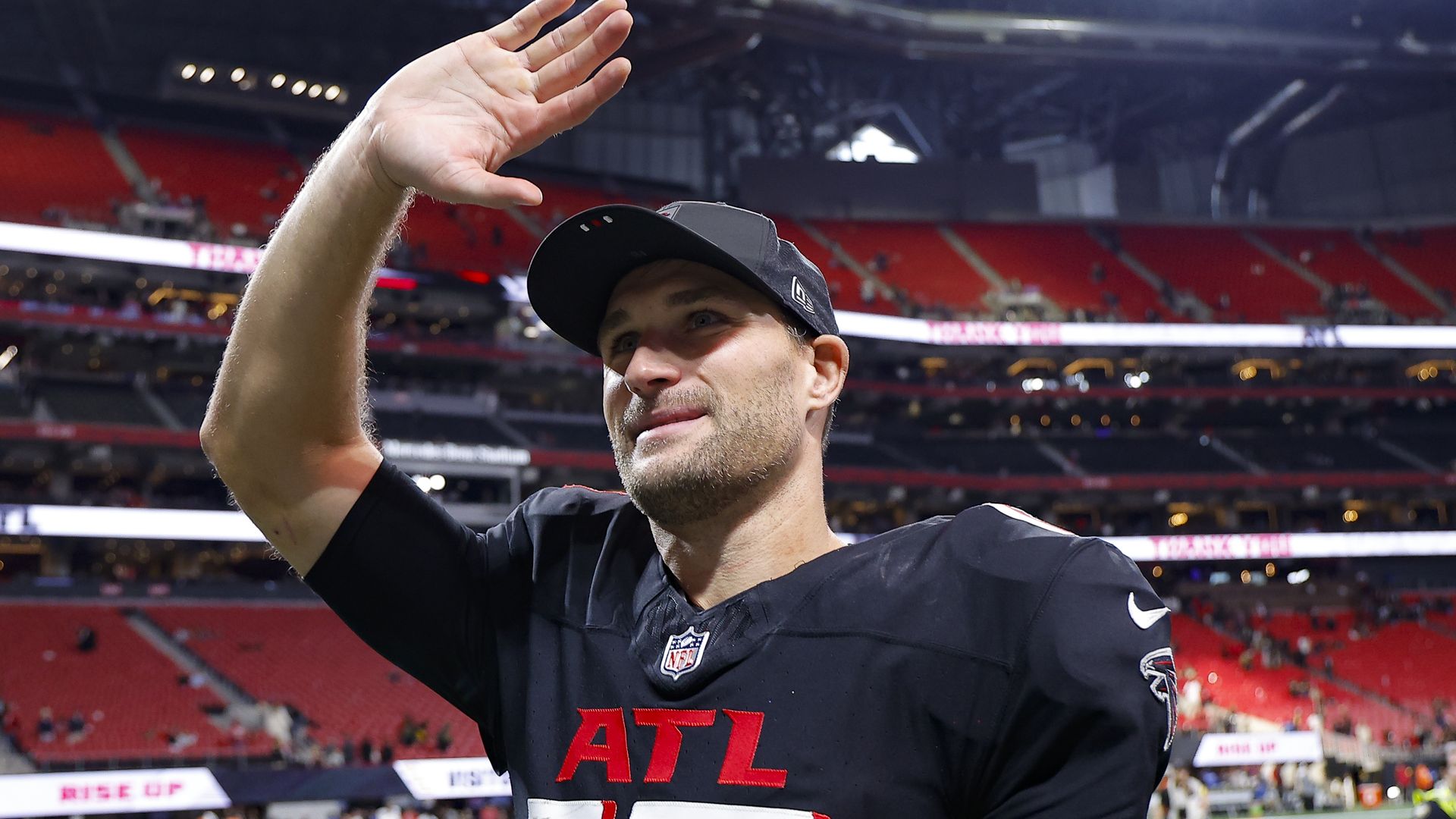 A football player wearing a black baseball hat and a black ATL jersey waves toward the crowd in a nearly empty stadium.
