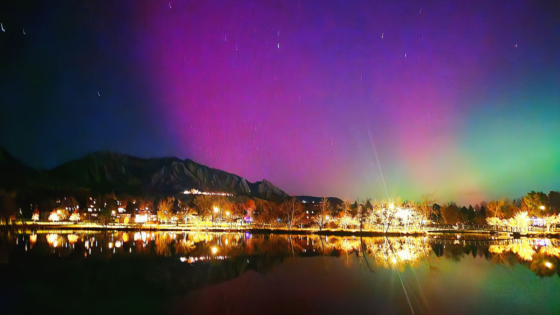 Night scene of a lake reflecting bright city lights, bare trees, and mountains under a sky with visible stars and colorful northern lights in purple, green, and blue hues.