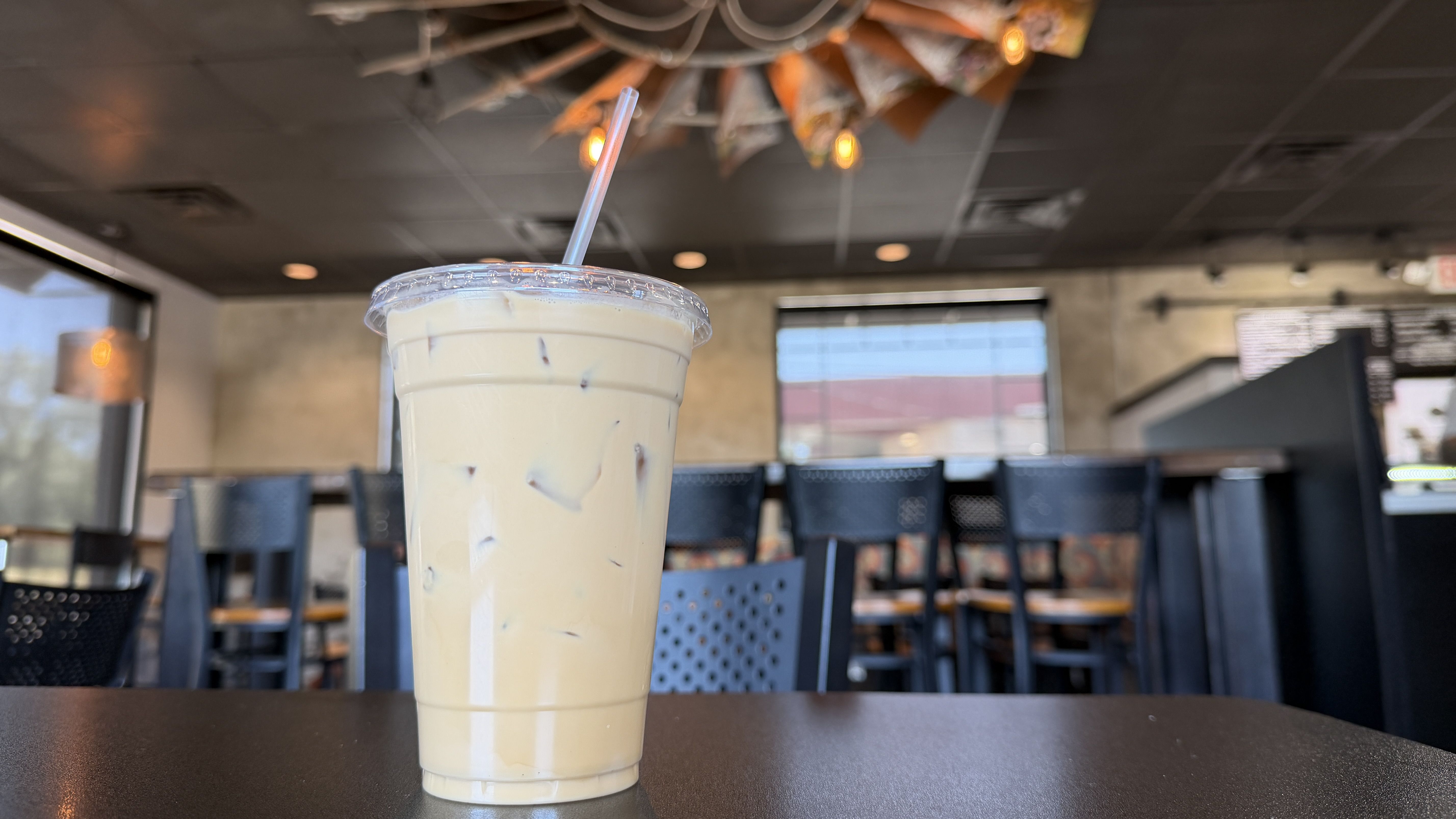 A tall iced coffee in a clear cup with a plastic lid and straw sits on a dark table inside a modern cafe; blurred chairs, windows, and warm ceiling lights in the background.