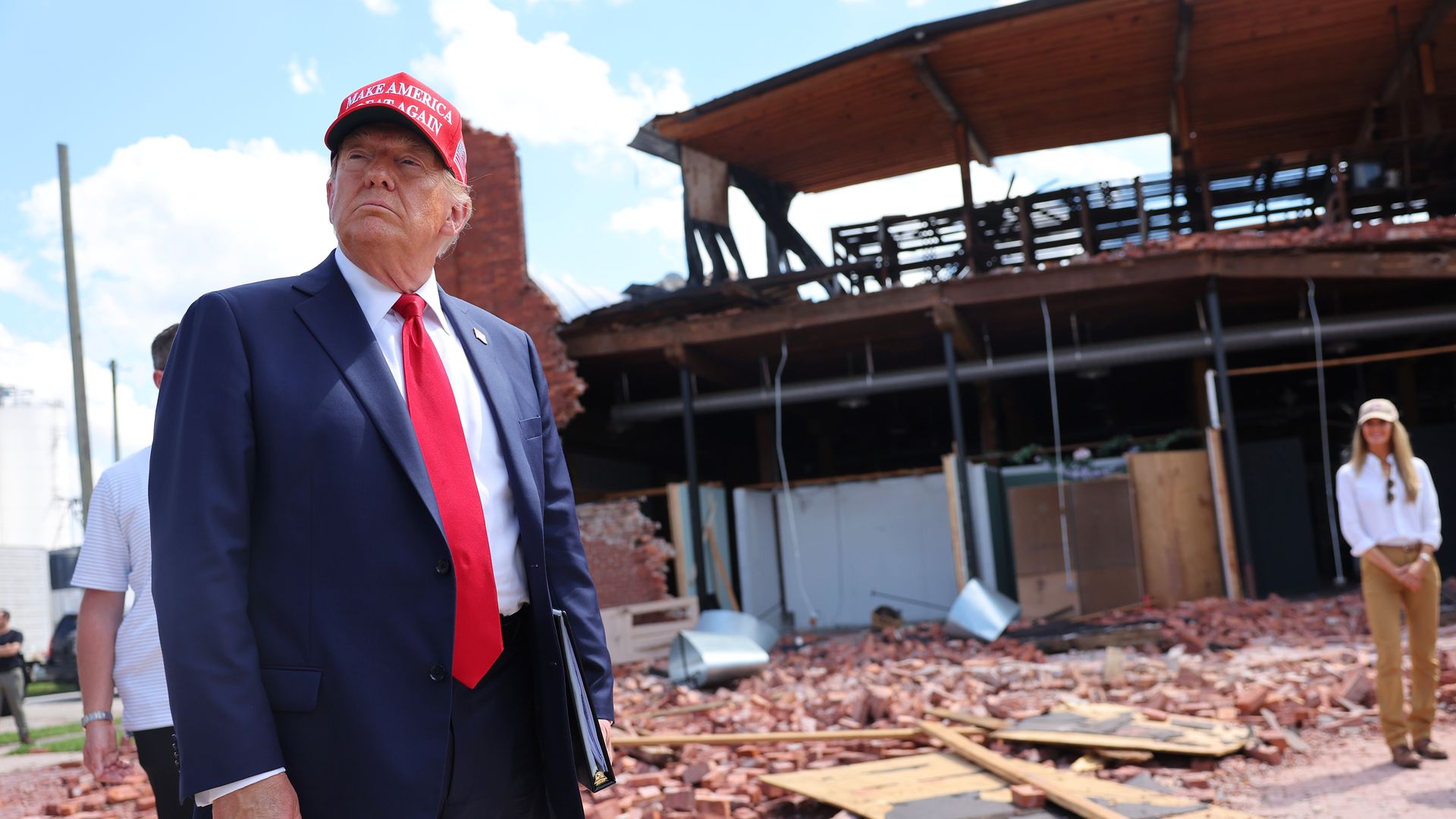 Donald Trump, in a blue suit, red tie and white shirt, stands in front of a damaged building.