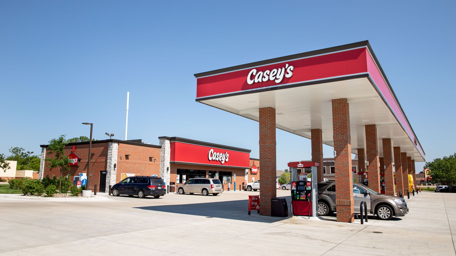 Wide view of a Casey's gas station with red sign, brick columns, white canopy, and clear blue sky. Several cars at fueling bays; storefronts visible in the background.