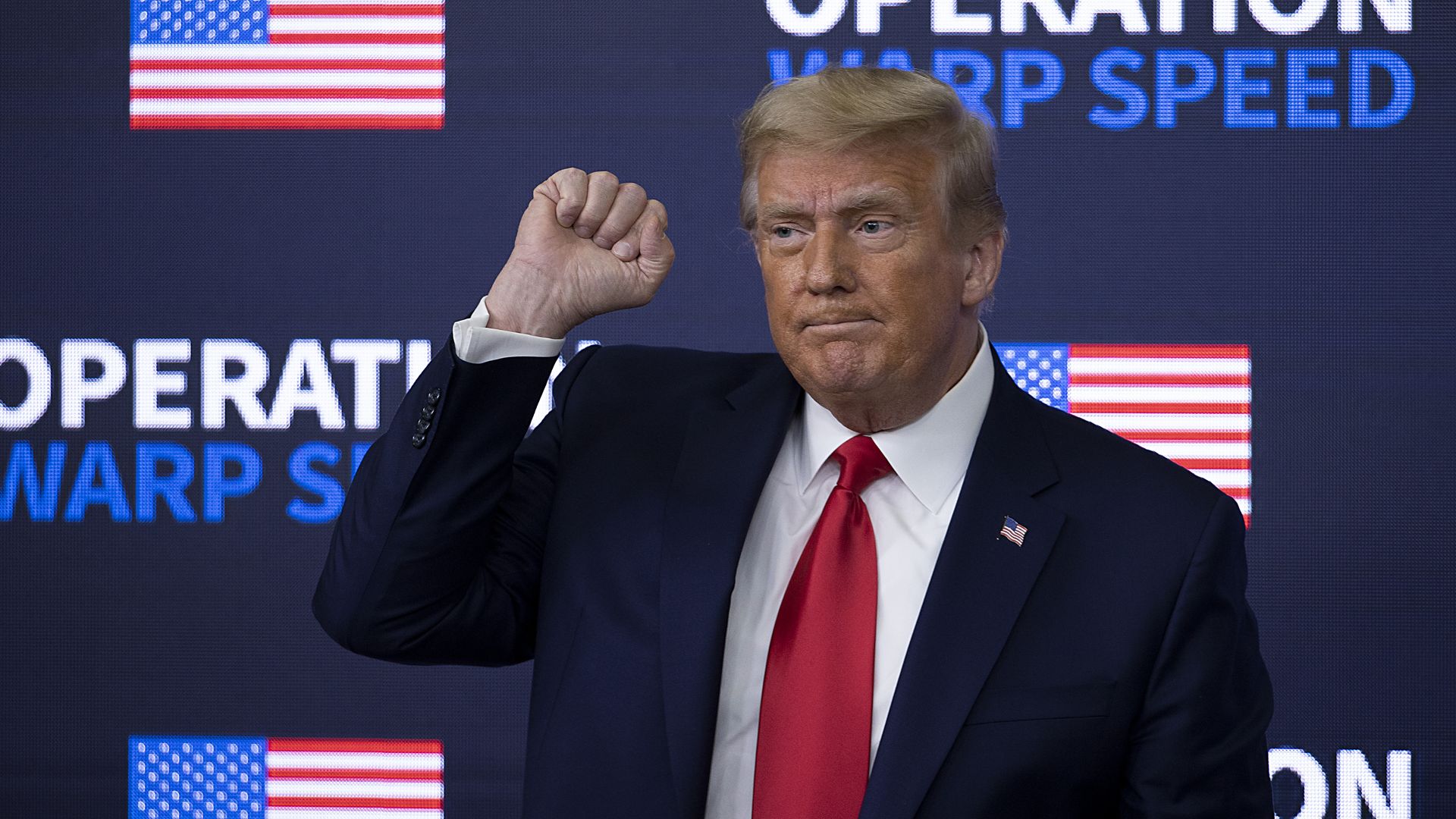 Trump holds a fist in the air while standing in front of a sign that reads "operation warp speed"