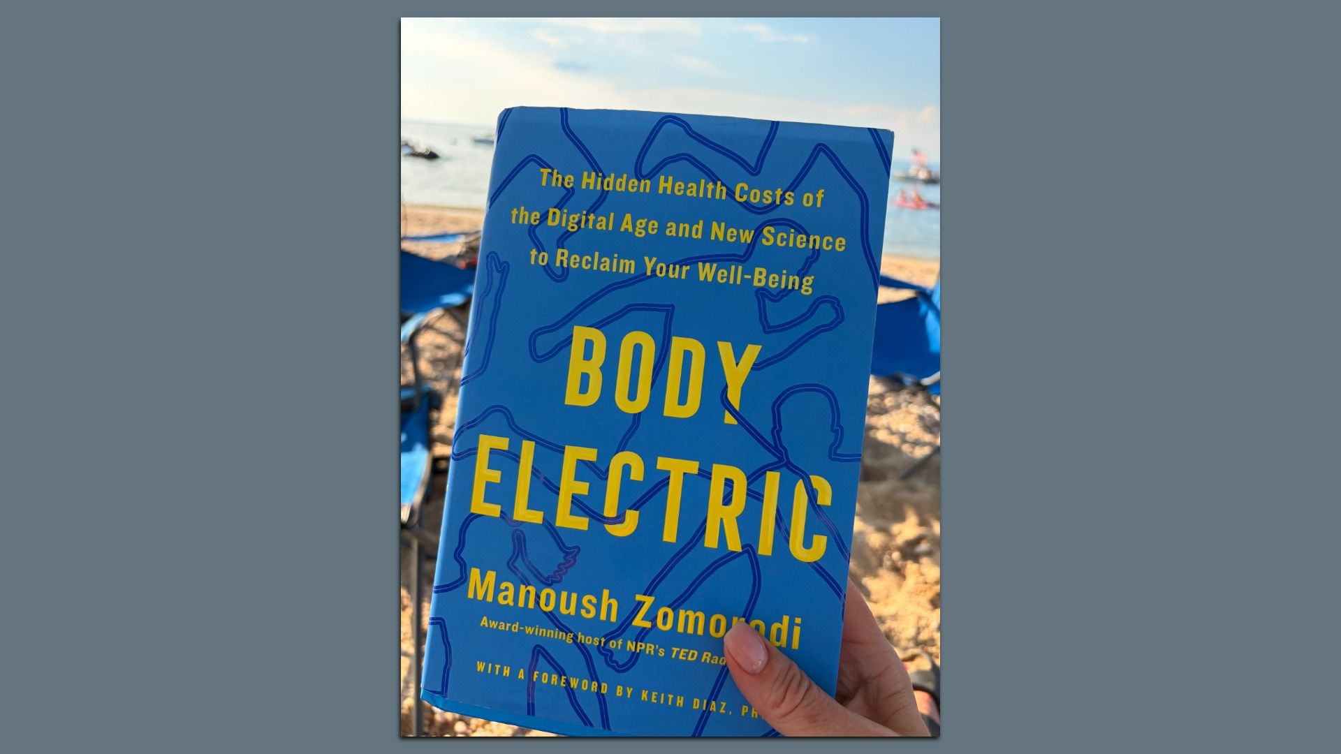 Blue book cover reading "Body Electric" in bold yellow letters with abstract blue outlines, held by a hand at a sunny beach; background shows sand, blue umbrellas, and the sea.