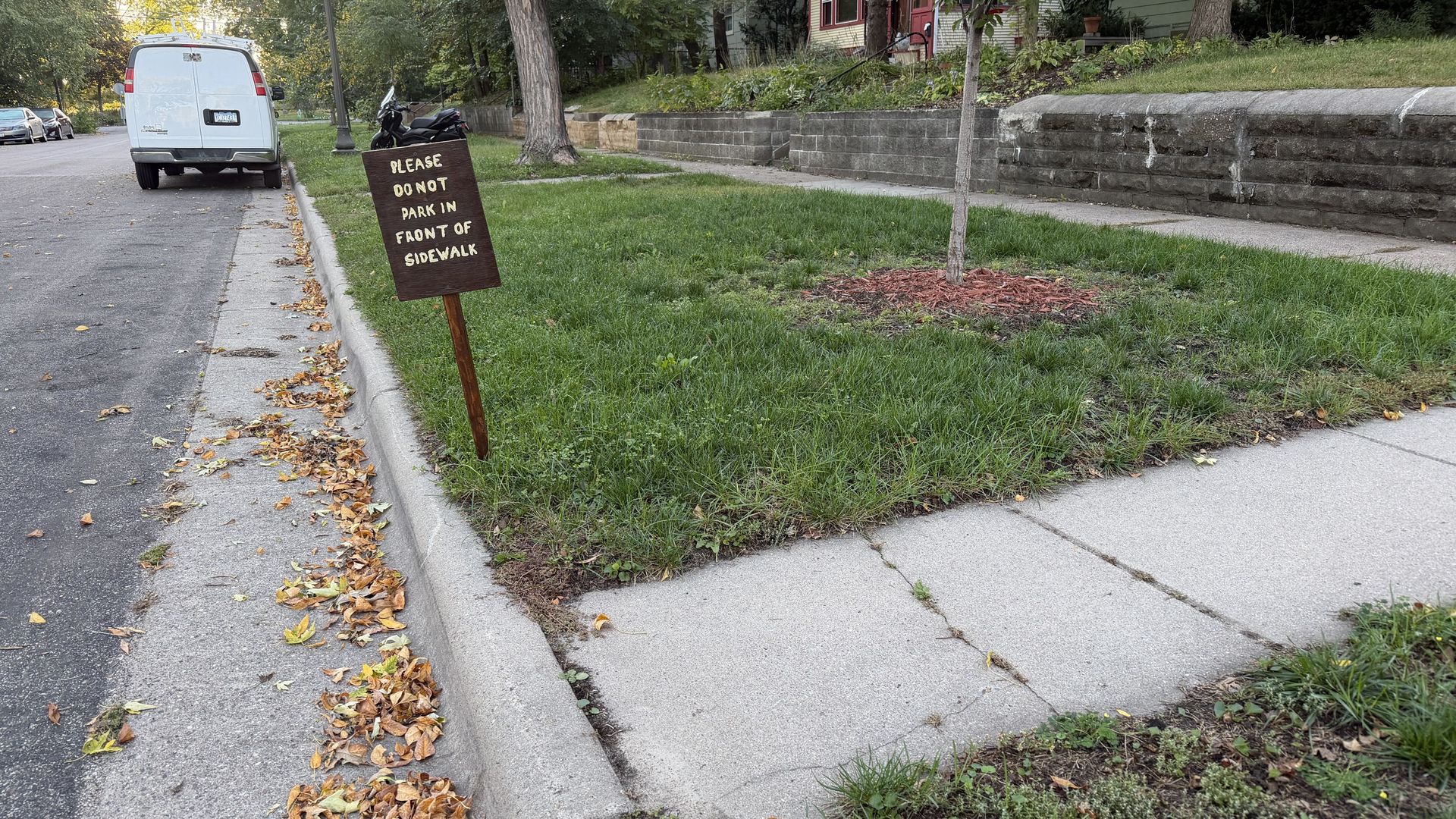 Residential street scene with a white van on the left. A brown sign leaning on a stake reads "PLEASE DO NOT PARK IN FRONT OF SIDEWALK." Leaves along the curb; trees and a brick wall in view.