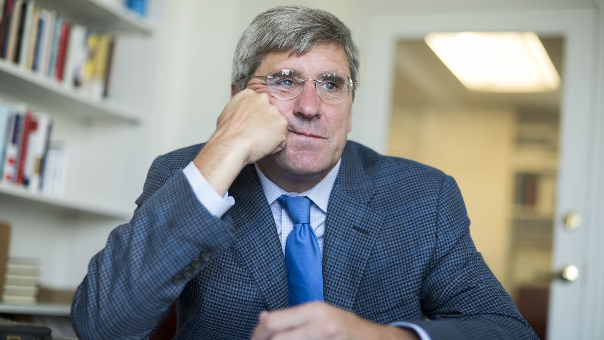 In this image, Stephen Moore sits at a desk and leans his face against one hand. He's wearing glasses. 