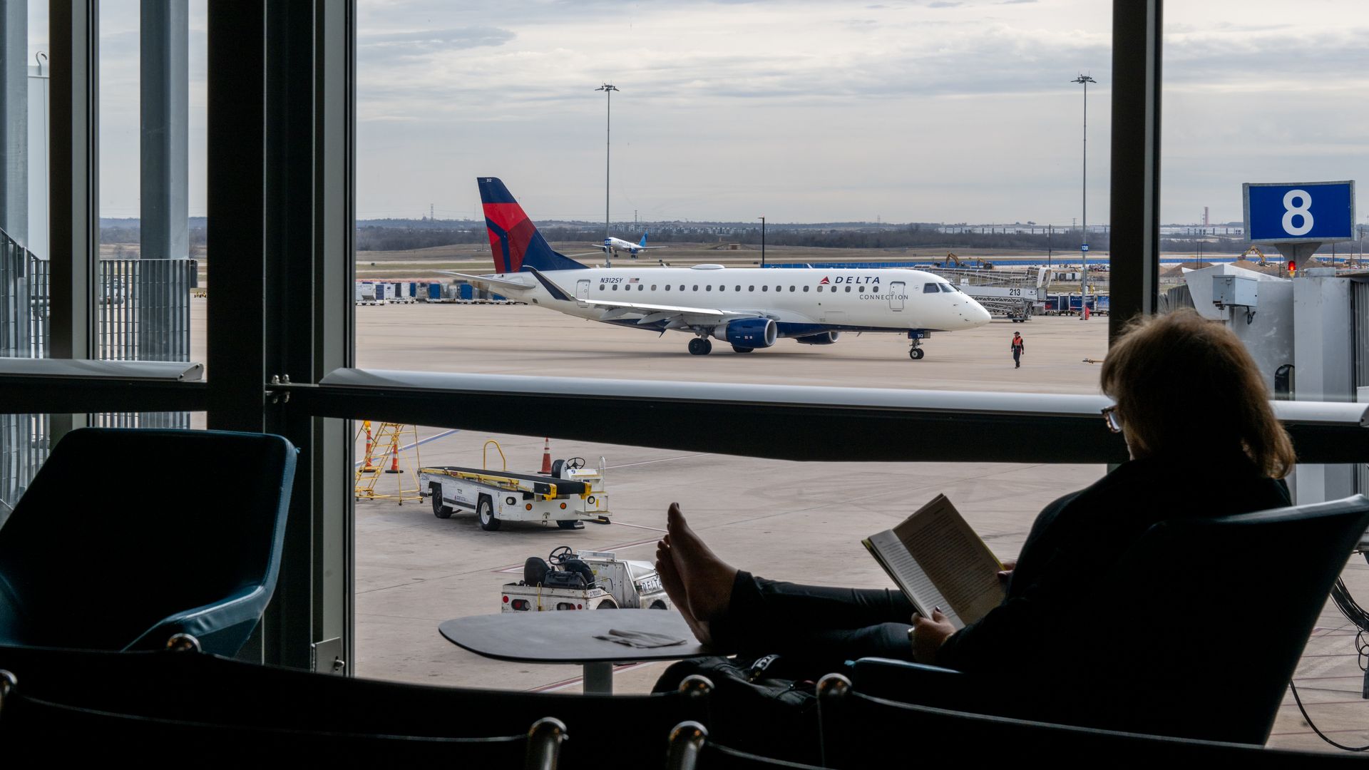 View from a terminal gate showing a Delta airplane on the tarmac near gate 8, framed by large windows; ground equipment and a passenger reading a book nearby.