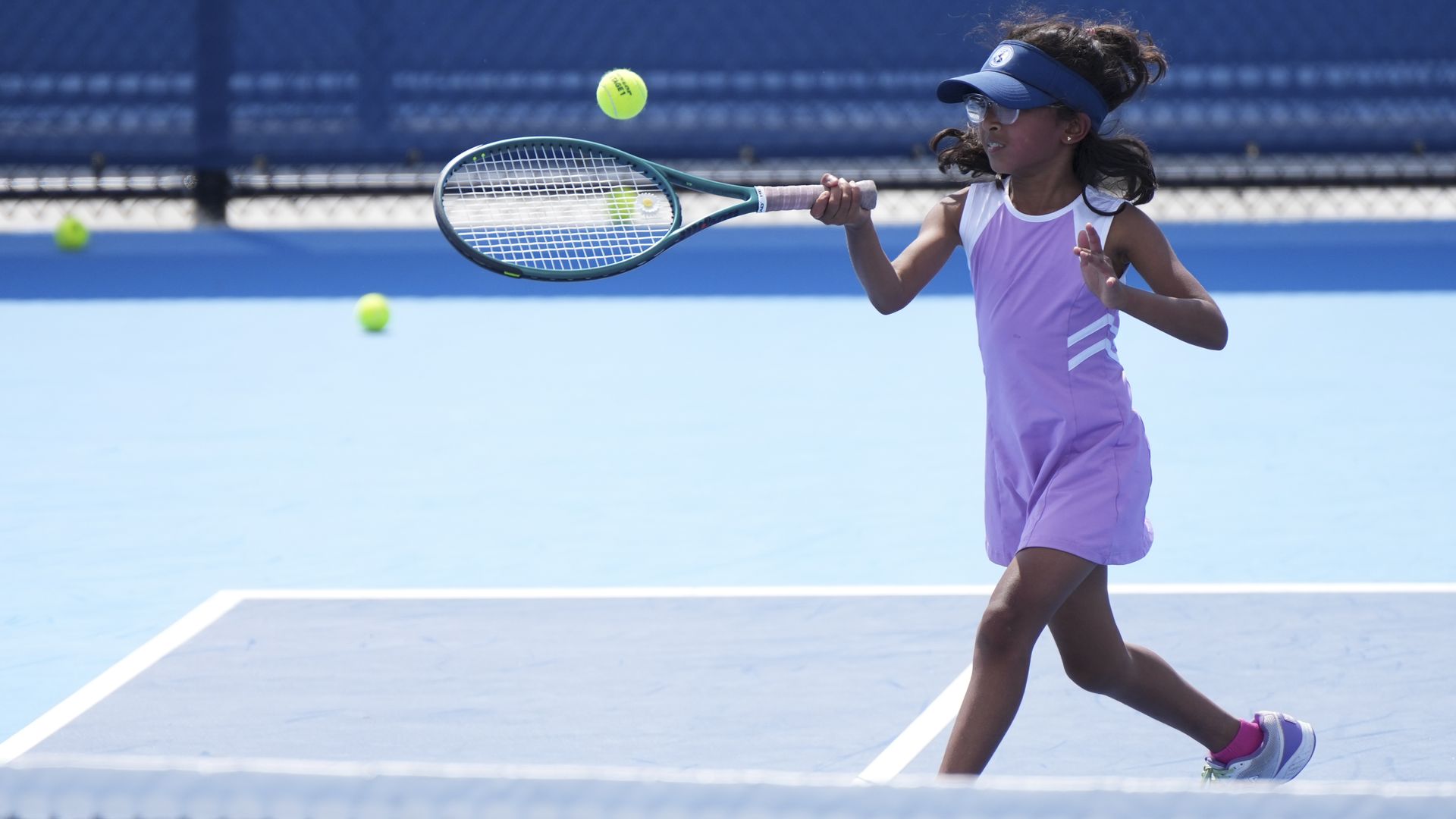 Young girl in a purple dress and blue visor hits a tennis ball with a racket on a bright blue court; neon green ball in midair, chain-link fence blurred in the background.