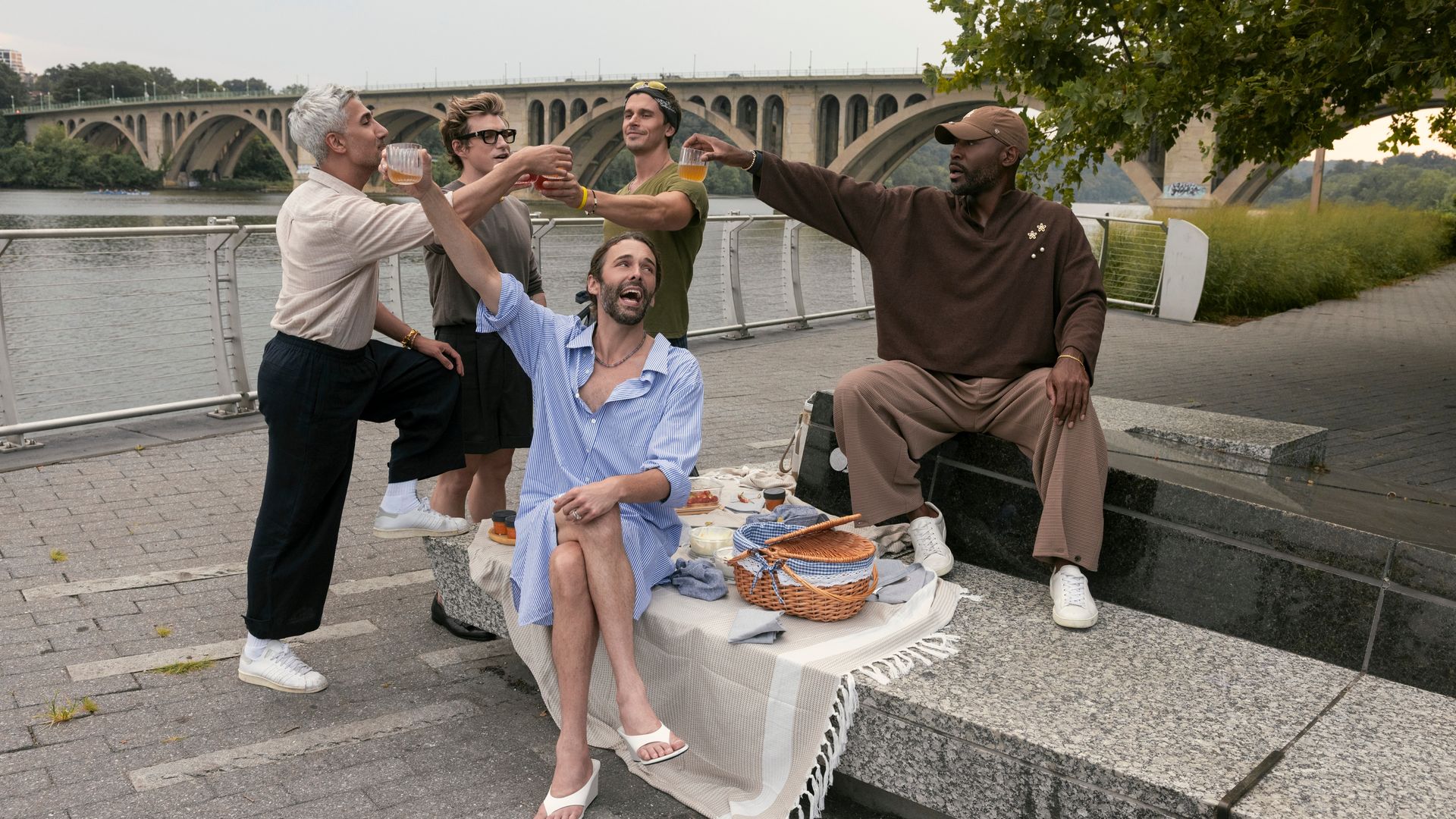 Five diverse friends outdoors near a river and stone bridge, gathering for a picnic with a basket. They cheer with drinks, smiling under a tree by the paved walkway.