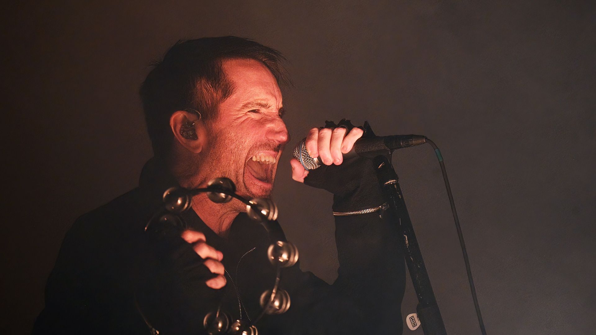 Trent Reznor of Nine Inch Nails performs on a dark stage, screaming into a mic while holding a tambourine. 