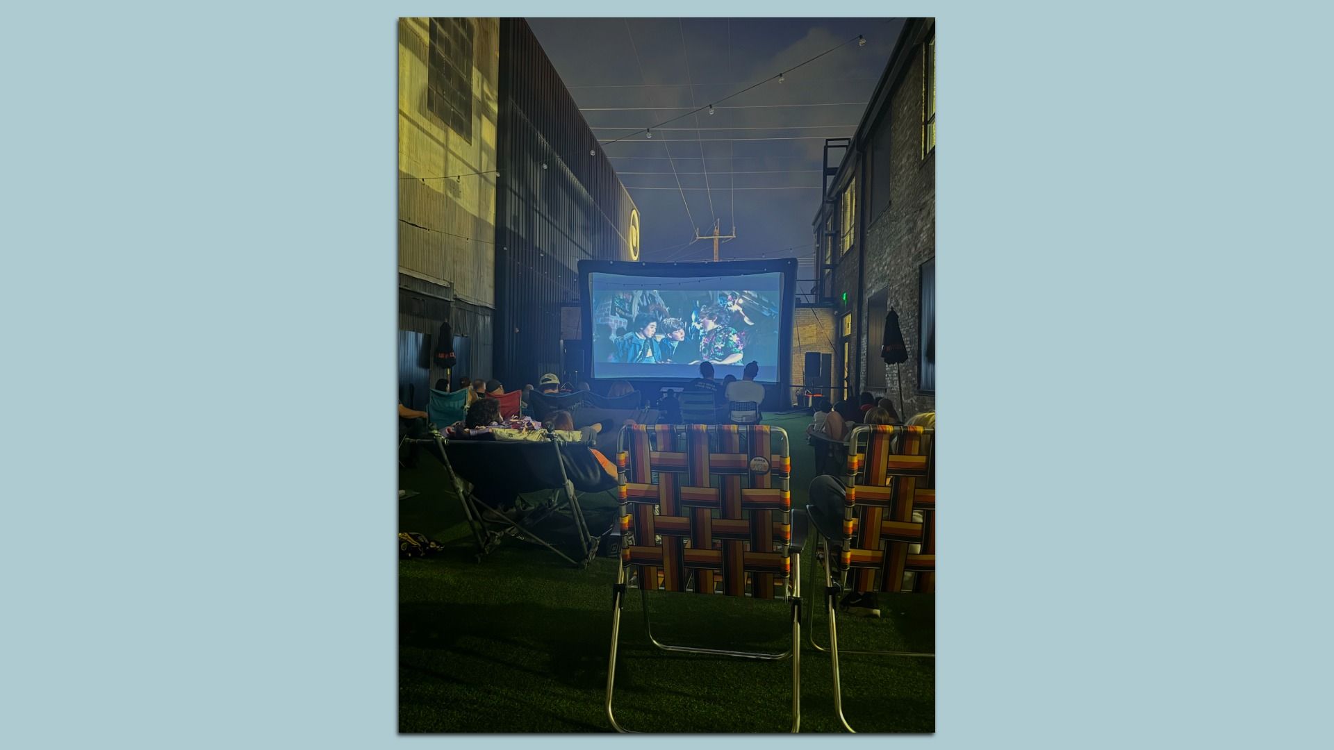 Outdoor movie night in an alley: a large screen between brick buildings, string lights overhead, and rows of colorful folding chairs and blankets as people watch a film.