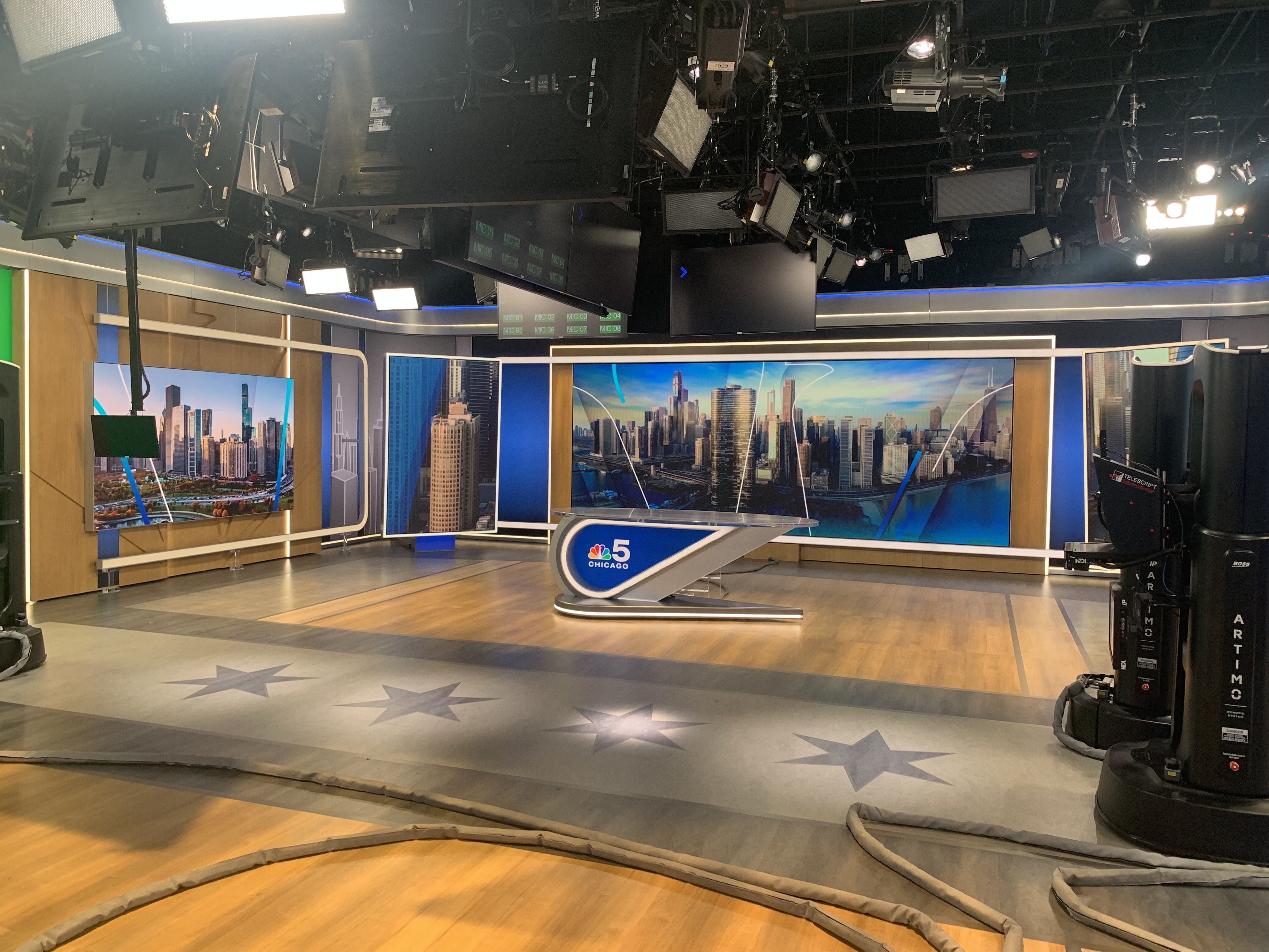 News studio with a curved desk displaying the NBC 5 Chicago logo, large cityscape screens, blue accents, and overhead lights; visible cameras and cables on a wood and gray stage.
