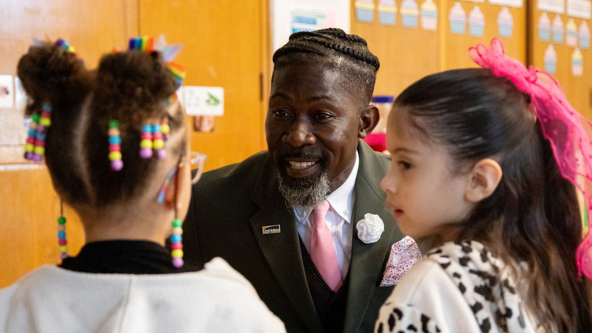 Smiling man in dark green suit and pink tie talks to two young girls, one with colorful beaded hair and the other with a pink bow, in a classroom setting with wooden cabinets.
