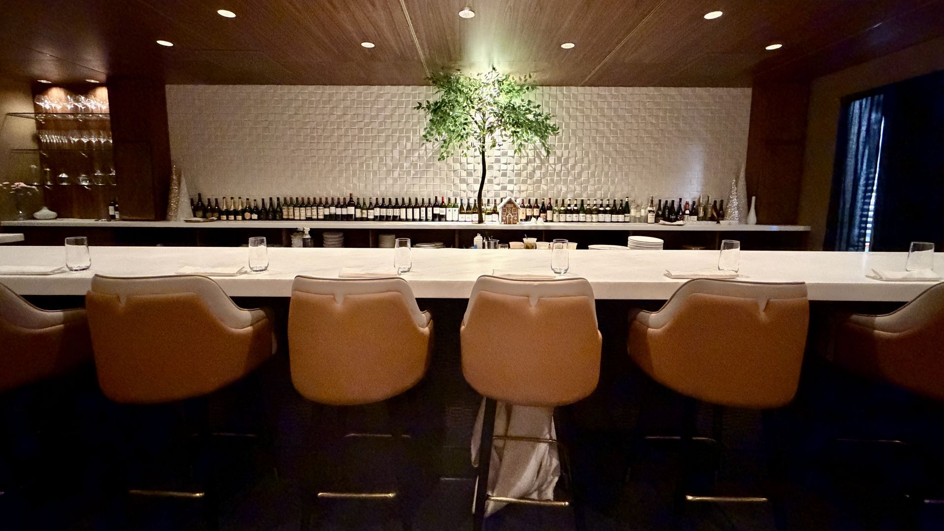 Elegant bar with six tan leather stools, white countertop, backlit textured wall, numerous bottles, and a small tree centerpiece under warm ceiling lights.