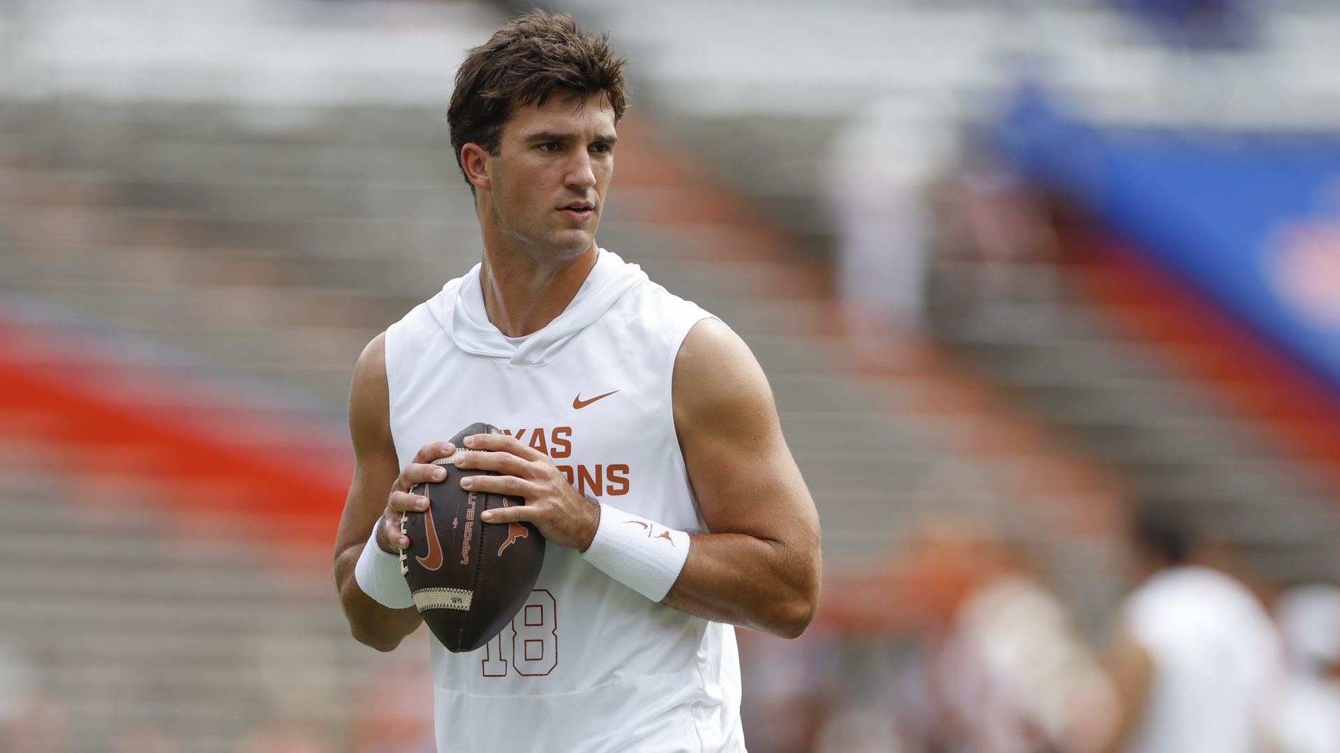 A football player in a white sleeveless Texas Longhorns shirt and black shorts holds a football, preparing to throw on a field with blurred stands in the background.