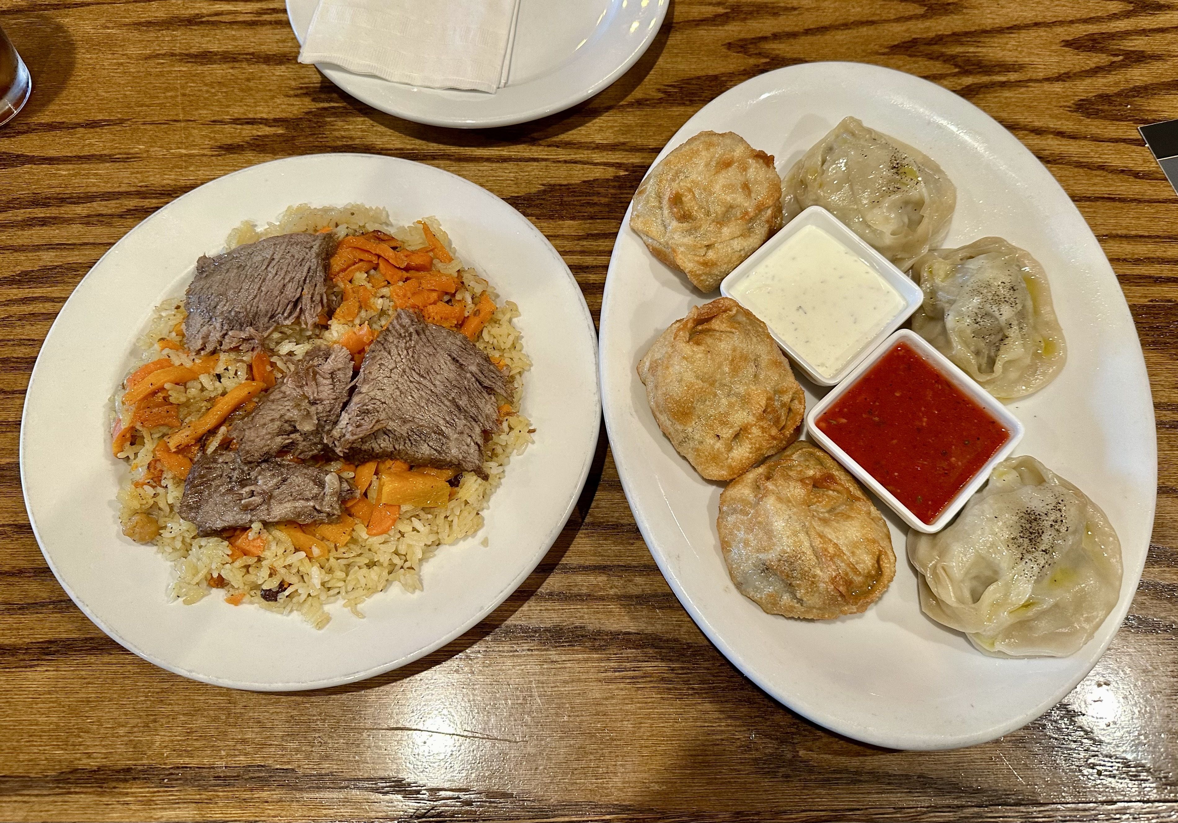 Two white plates on a wooden table: left plate has beef and carrot fried rice with beef slices; right plate features dumplings with red and white dipping sauces and several fried pastries.