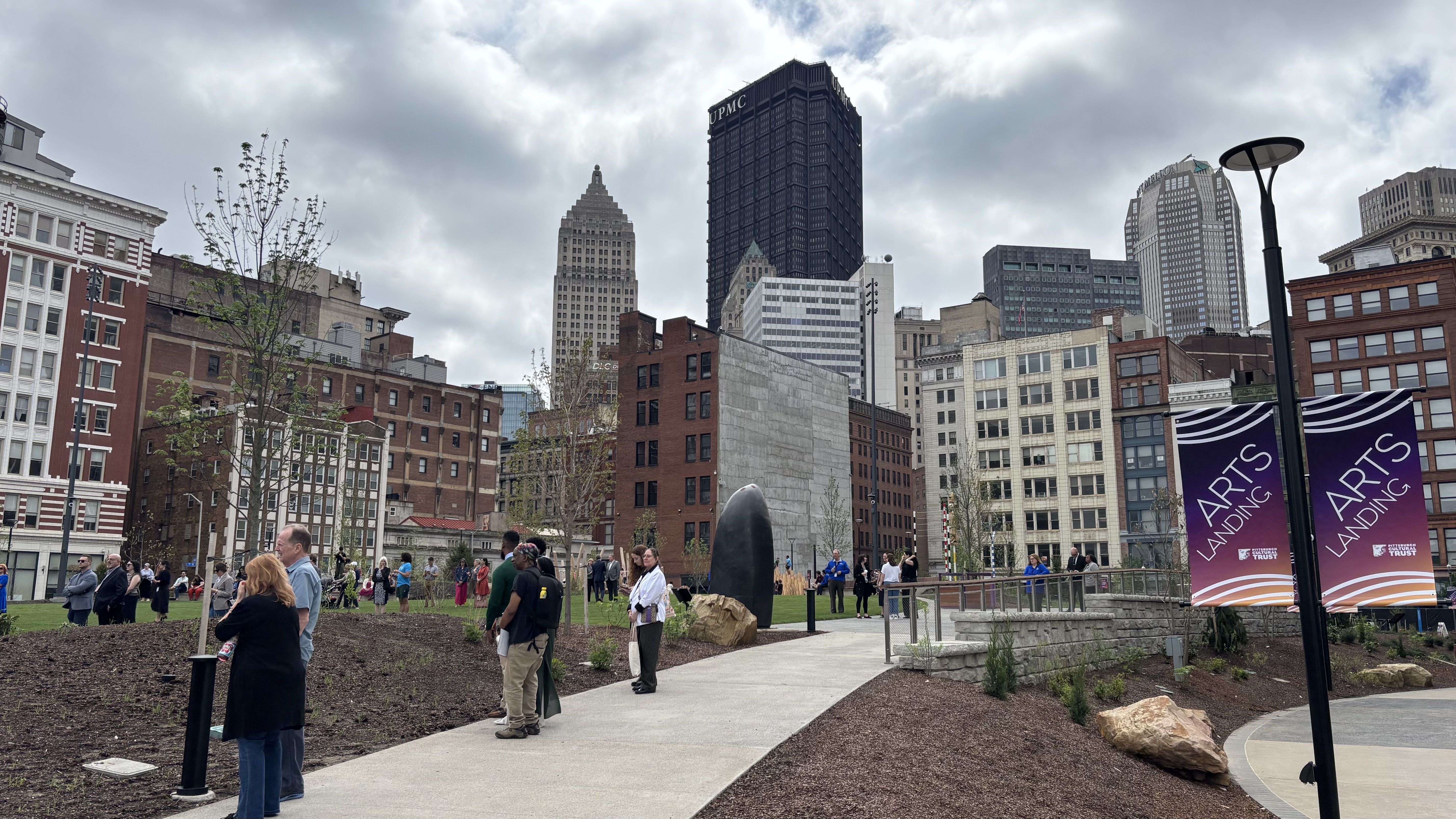 City park scene with people walking on a paved path beneath a cloudy sky. In the skyline, tall UPMC and other buildings rise; a dark oval sculpture sits by the path near purple "ARTS LANDING" banners.