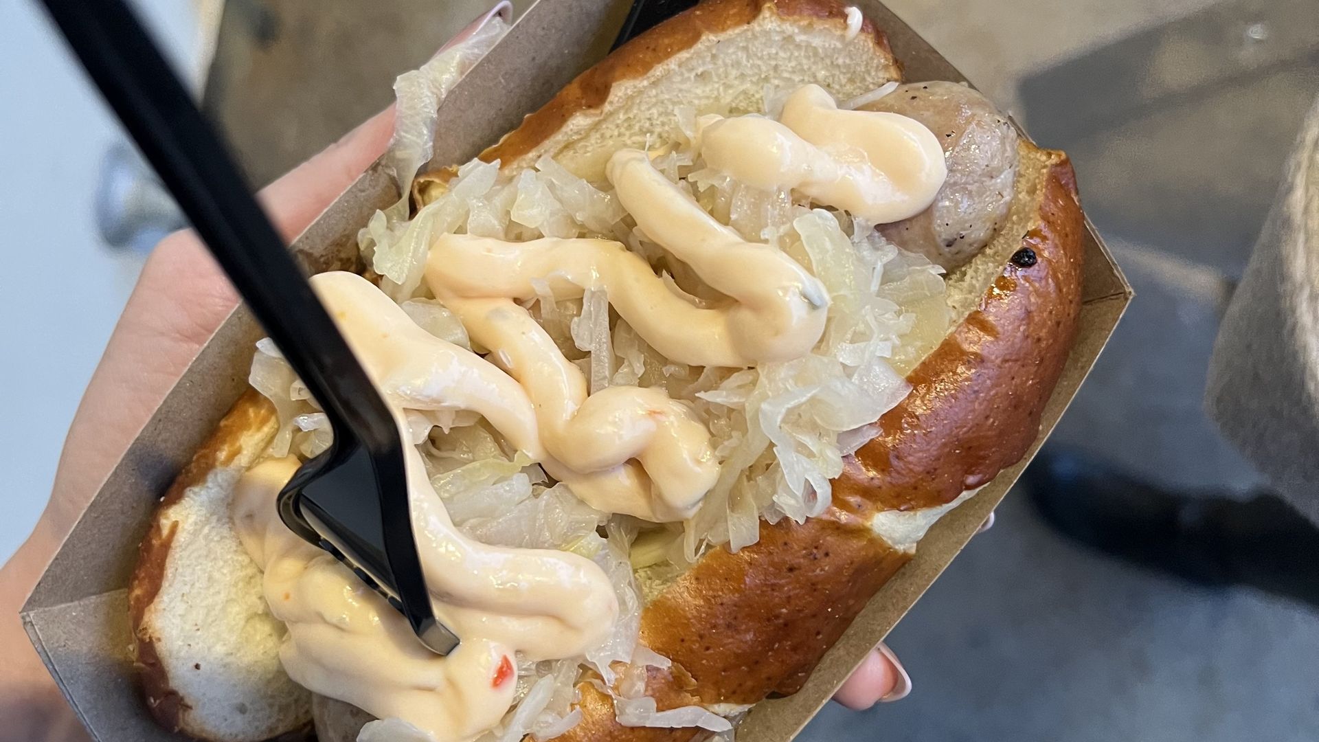 A hand holding a sausage in a soft brown pretzel bun topped with sauerkraut and a zigzag of cream-colored cheese sauce, served in a brown paper tray with a black plastic fork on top.
