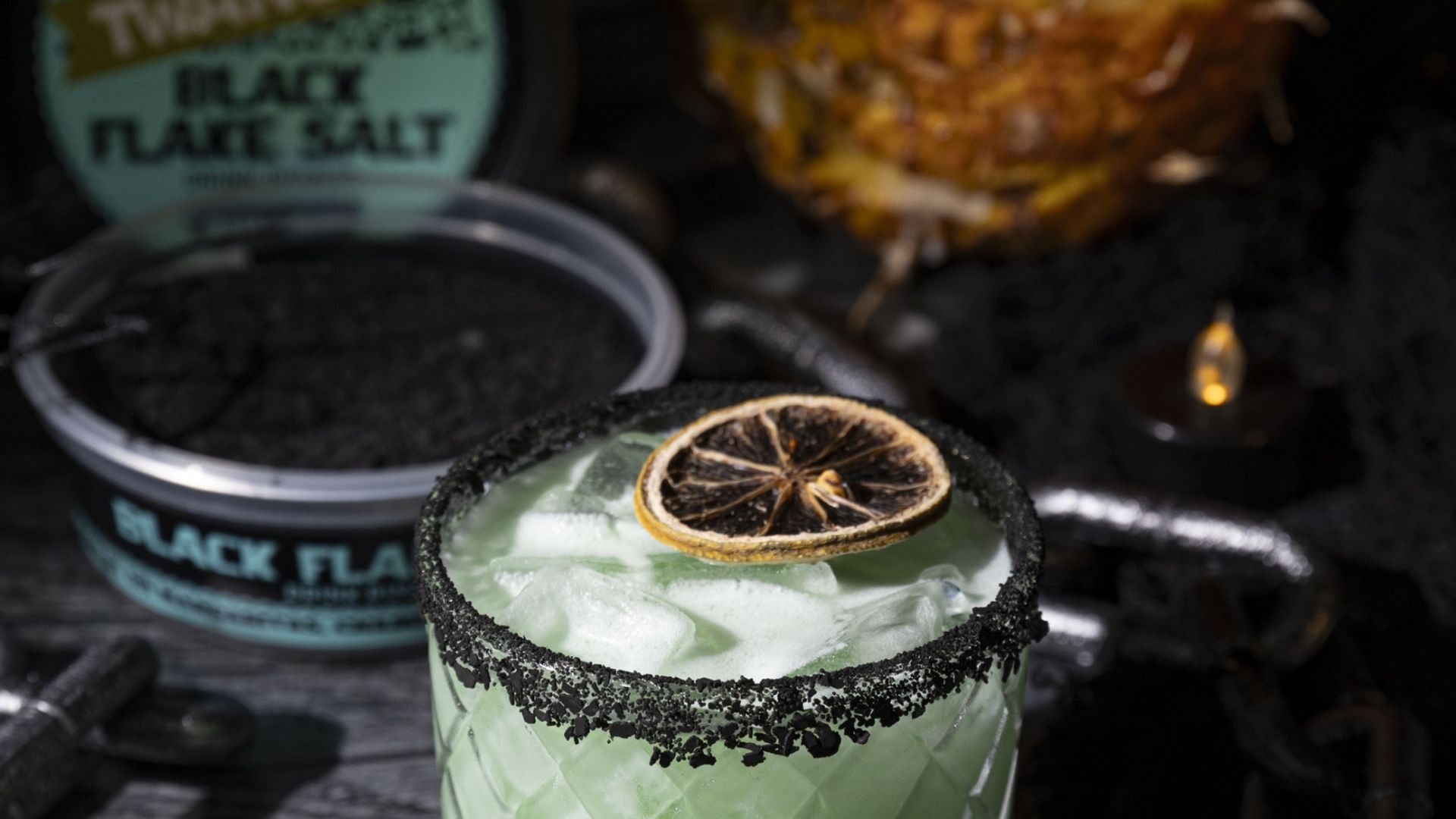 A cocktail that is colored light green in a glass with ice rimmed with black flake salt and topped with a dried citrus slice. It sits against a dark background with a container of black flake salt behind it.