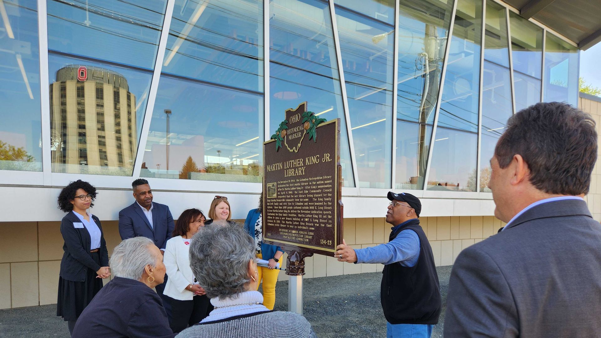 A man reads text of a historical marker outside a library to a crowd. 