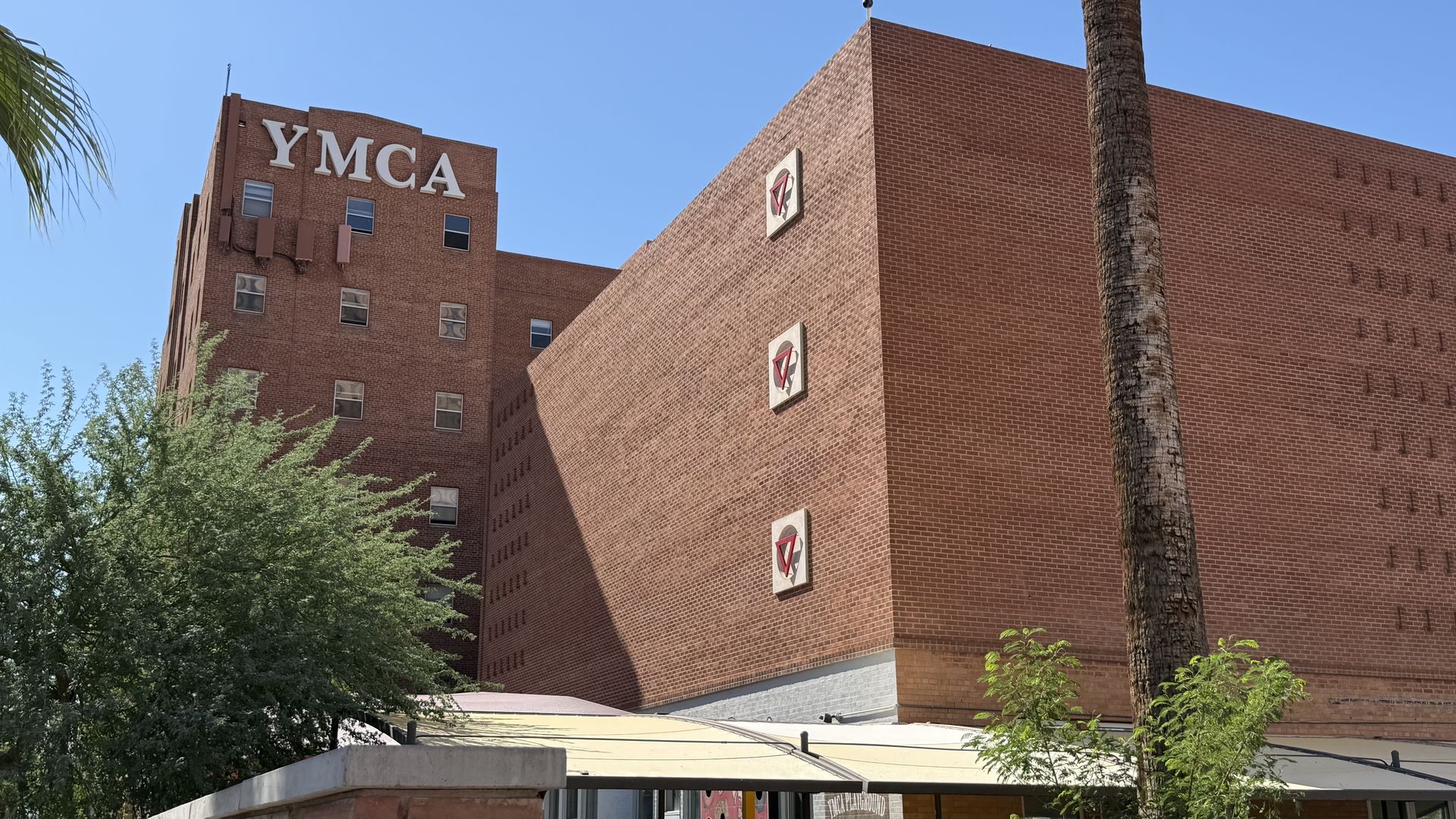 Brick YMCA building under a clear blue sky, featuring a large white YMCA sign on the left. A tall palm trunk on the right, with green foreground trees and small square logo panels on the brick wall.
