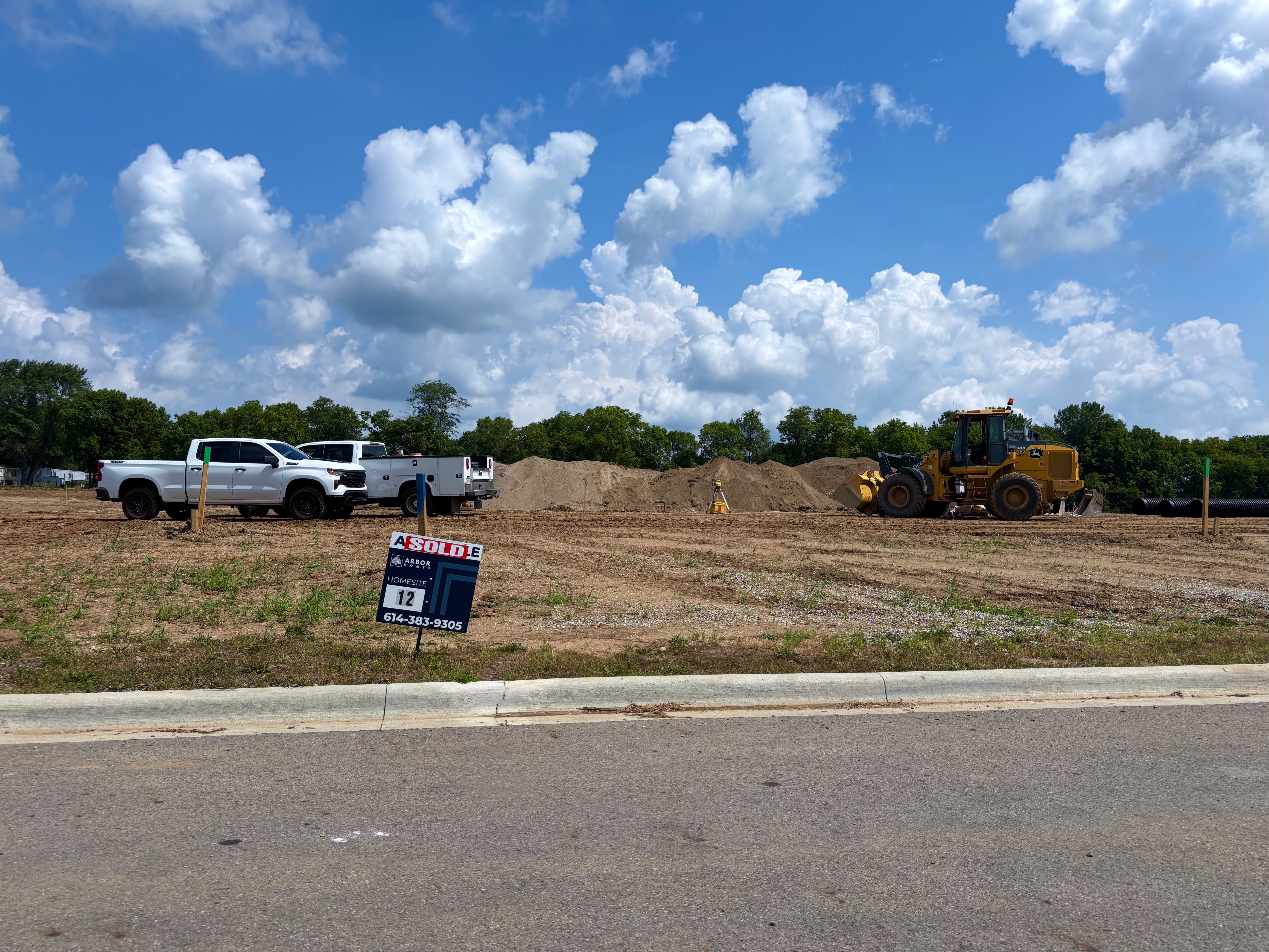 Construction site under a bright blue sky with white clouds. In the foreground are white trucks and a yellow bulldozer, piles of dirt, and a "SOLD" real estate sign with a phone number.