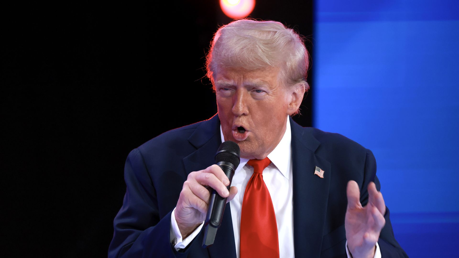 Donald Trump wearing a dark suit, white shirt and red tie holds a microphone as he speaks into it