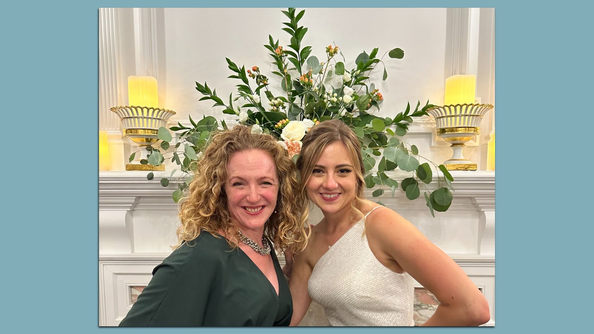 Two smiling women pose for a selfie in front of a white mantel with a lush greenery arrangement of leaves and white flowers, flanked by glowing amber candles on gold holders.