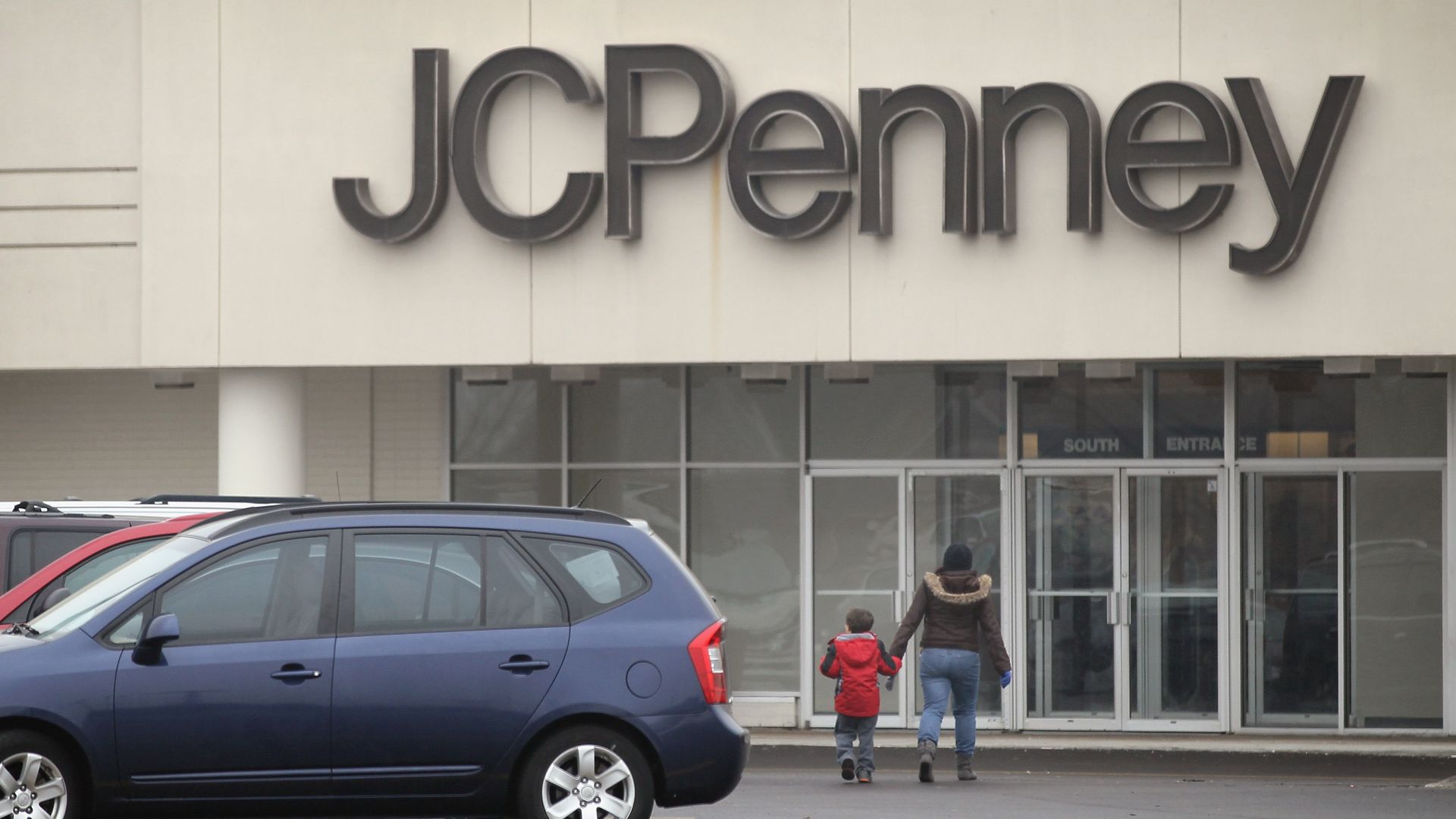 Front of a JC Penney store with a large dark sign on a pale facade. Two people walk toward glass doors marked South Entrance while several cars sit in the parking lot.