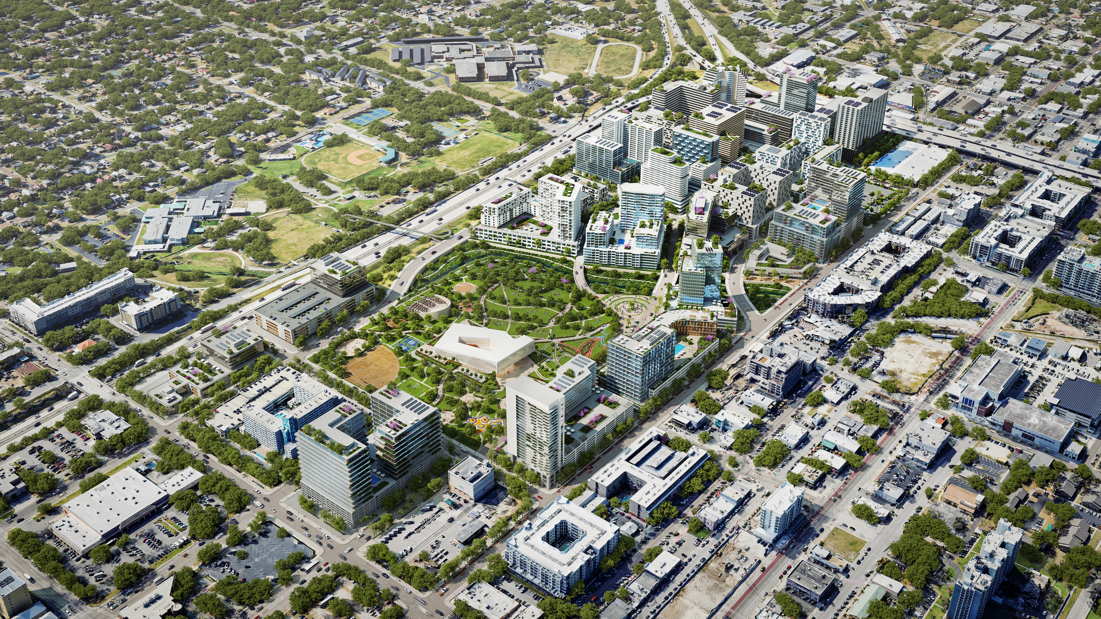 Aerial view of a modern urban area with mixed-use high-rise buildings surrounding a large park with green spaces, walking paths, and recreational areas, near a highway and residential neighborhoods.