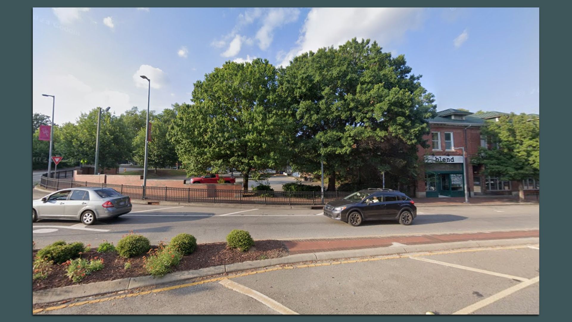 Street scene with two cars passing by; large green trees and a red brick building with a sign reading "blend" are visible under a blue sky with scattered clouds.