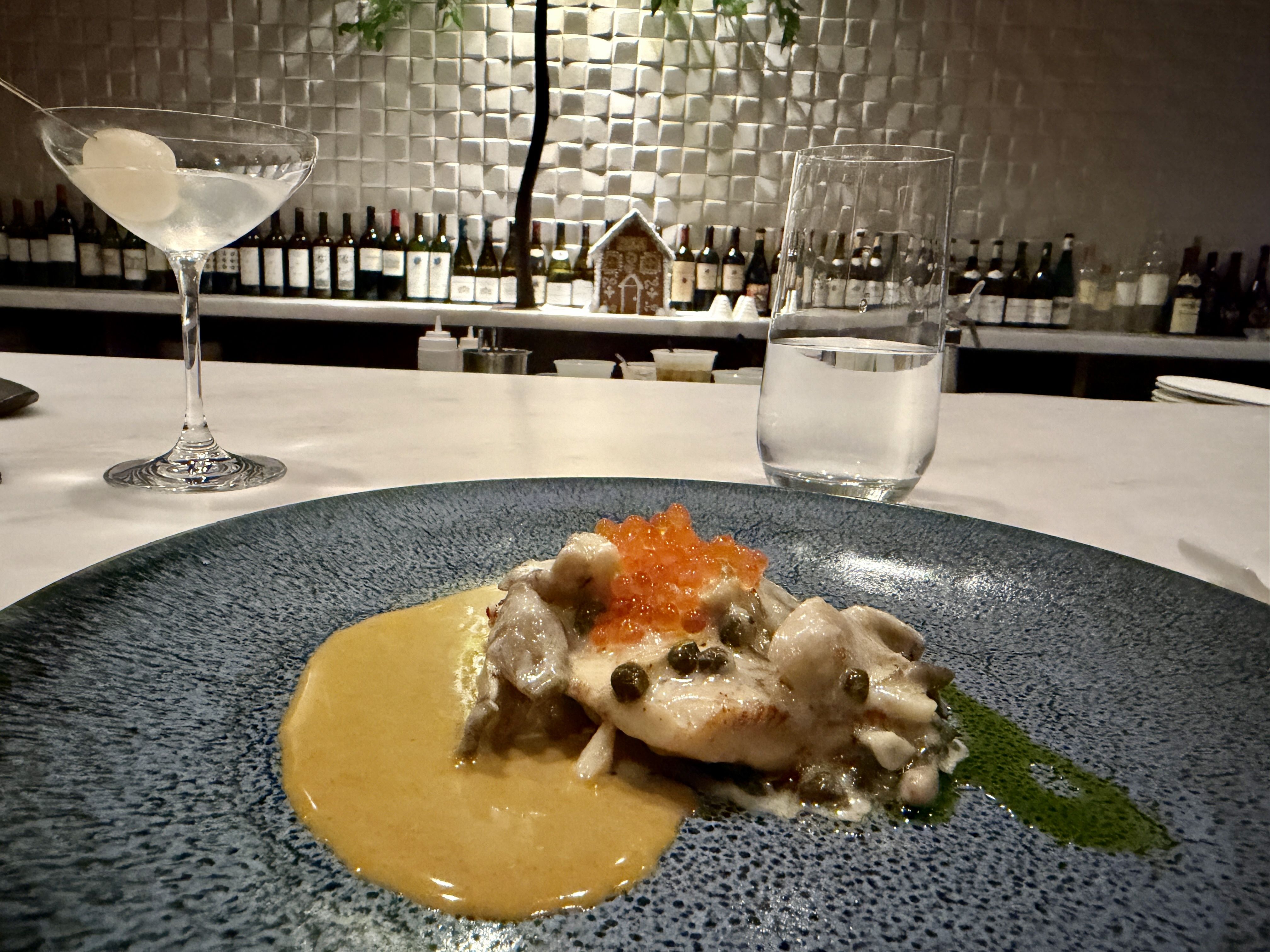 Blue plate with a gourmet dish featuring white fish topped with orange roe and capers in creamy sauce, served with a green herb drizzle, a martini glass with olive, and a water glass on a marble bar.