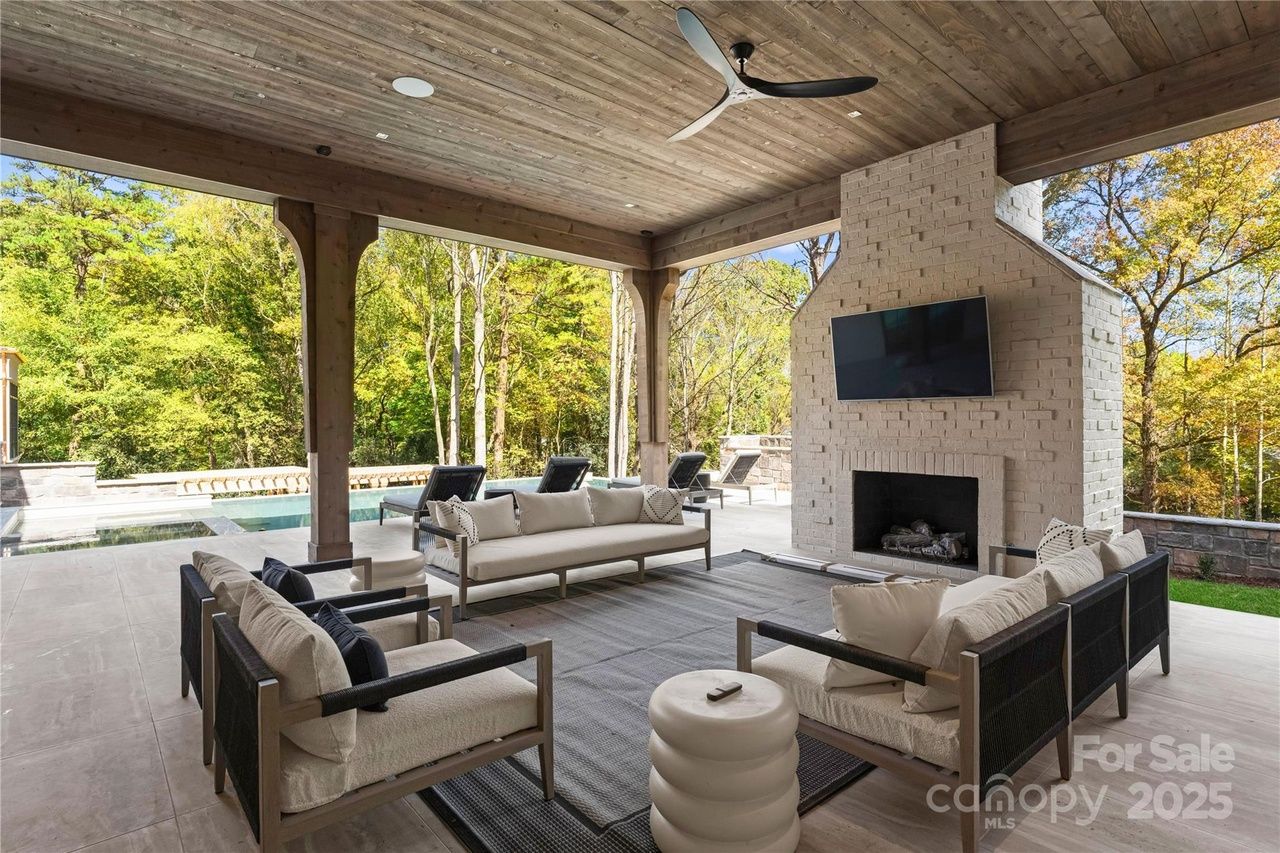 Covered outdoor living area with beige sofas, a gray rug, white brick fireplace with mounted TV, a ceiling fan, and a view of a pool and green trees in the background.
