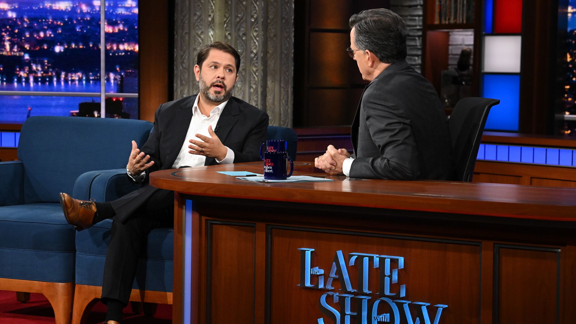 Two men in suits engaged in conversation on the set of "The Late Show," featuring a wooden desk with the show's logo, blue couch, and nighttime cityscape backdrop.
