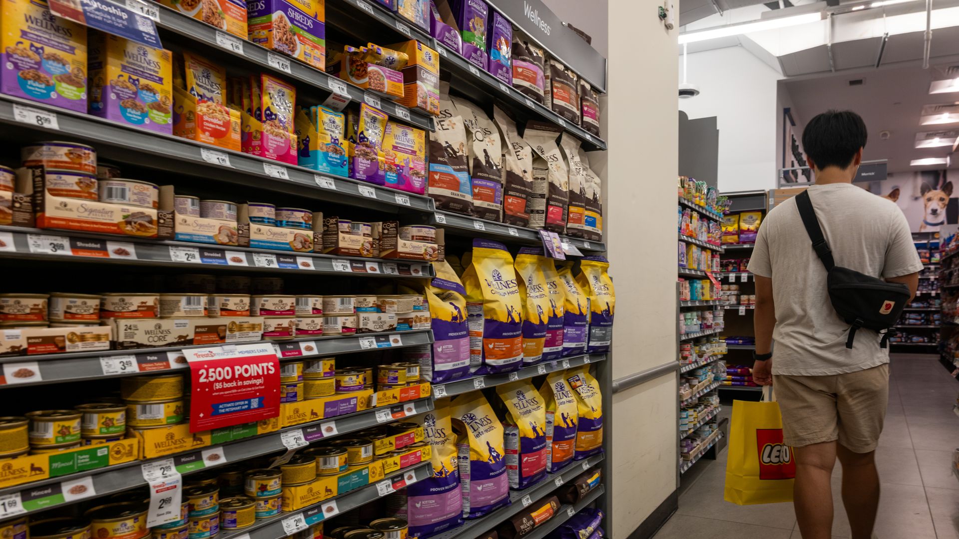 A person in a white shirt and beige shorts walks down a pet food aisle at a store, carrying a yellow LEGO shopping bag; shelves are stocked with colorful pet food bags and cans.