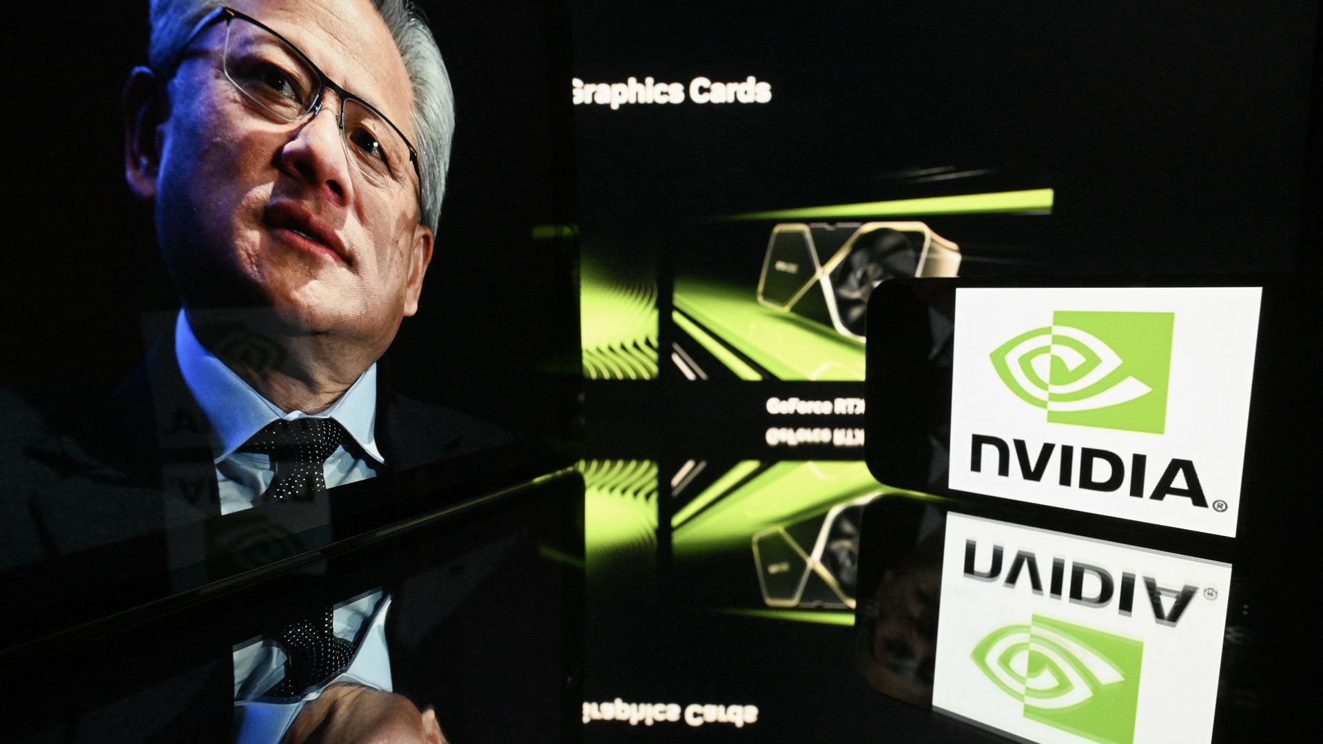 Man with glasses and gray hair wearing a suit and tie is reflected on a black surface next to an illuminated Nvidia logo and graphics card images with green and black colors.